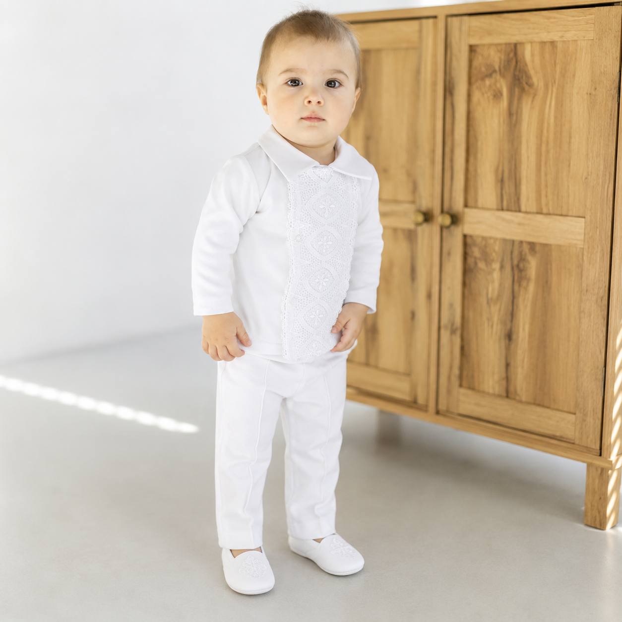 A toddler standing in a white traditional outfit with embroidered details on the chest, paired with white pants and white shoes, in front of a wooden cabinet.