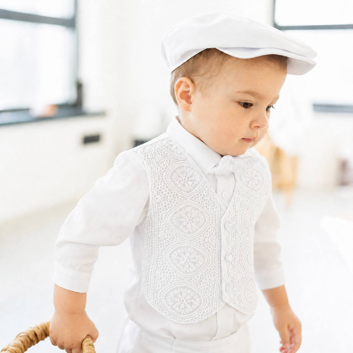 A toddler boy wearing a white long-sleeve shirt, a white lace vest, a white bow tie, and a white flat cap, walking while holding a wicker basket handle in a bright indoor setting.