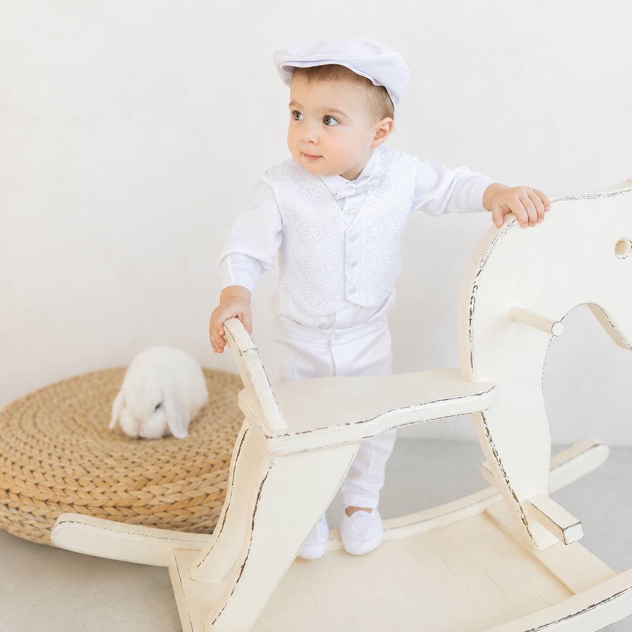 A baby boy wearing a white long-sleeve shirt, matching vest, trousers, and a white cap stands beside a large white rocking horse. A small white rabbit sits on a woven round mat in the background against a soft off-white studio wall.