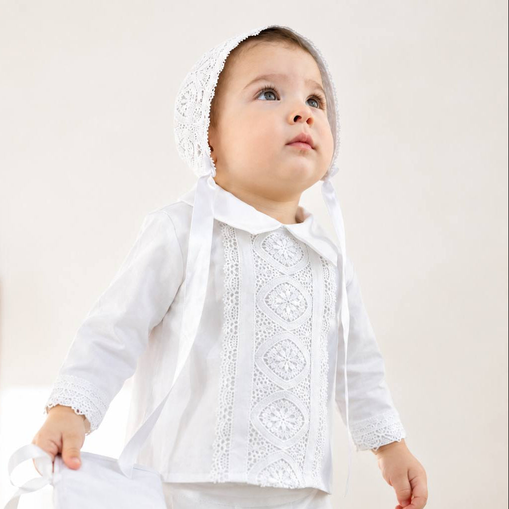 A toddler wearing a white lace and satin first communion outfit with a matching bonnet, standing in a softly lit indoor setting.