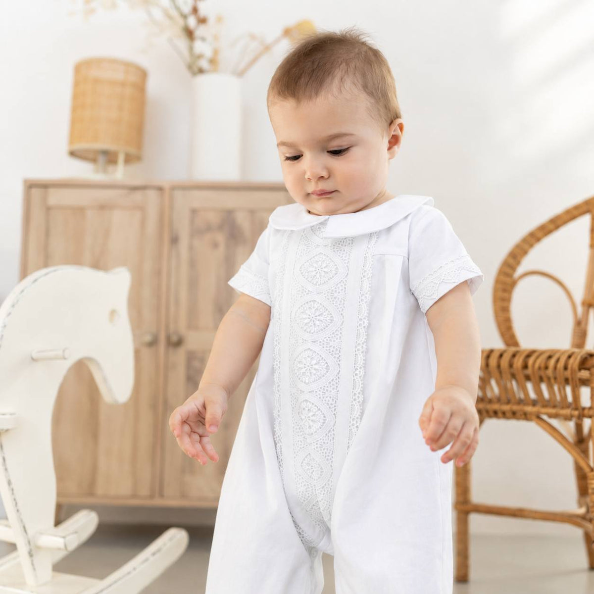 A baby wearing a white short-sleeve lace-trimmed romper or jumpsuit, standing in a bright, softly styled room with wooden furniture and a wicker chair in the background.
