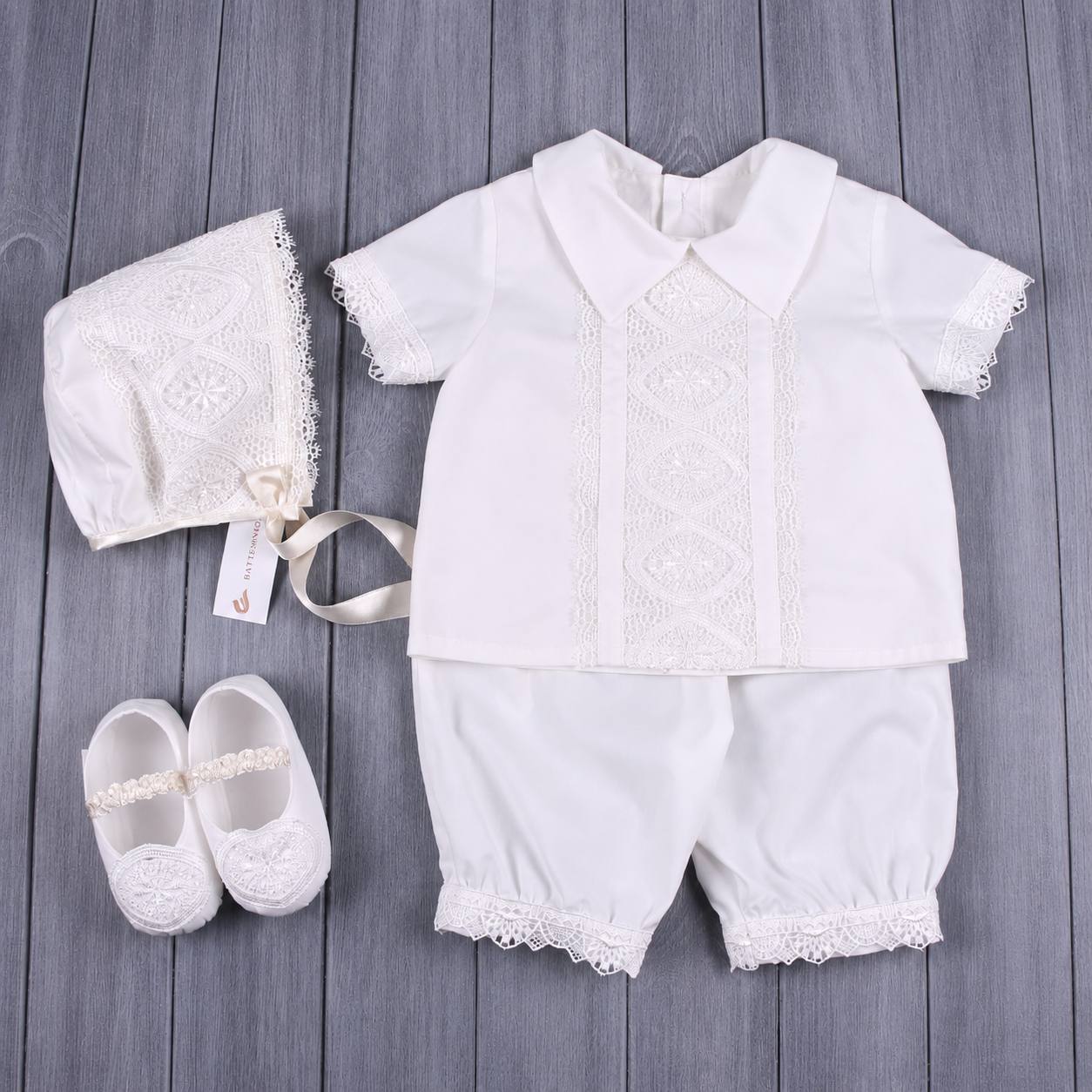 A baby christening outfit set laid flat on a gray wood plank background, including a white short-sleeve collared shirt with lace embroidery, matching white shorts with lace trim, a white lace bonnet with a ribbon, and a pair of white lace baby shoes.
