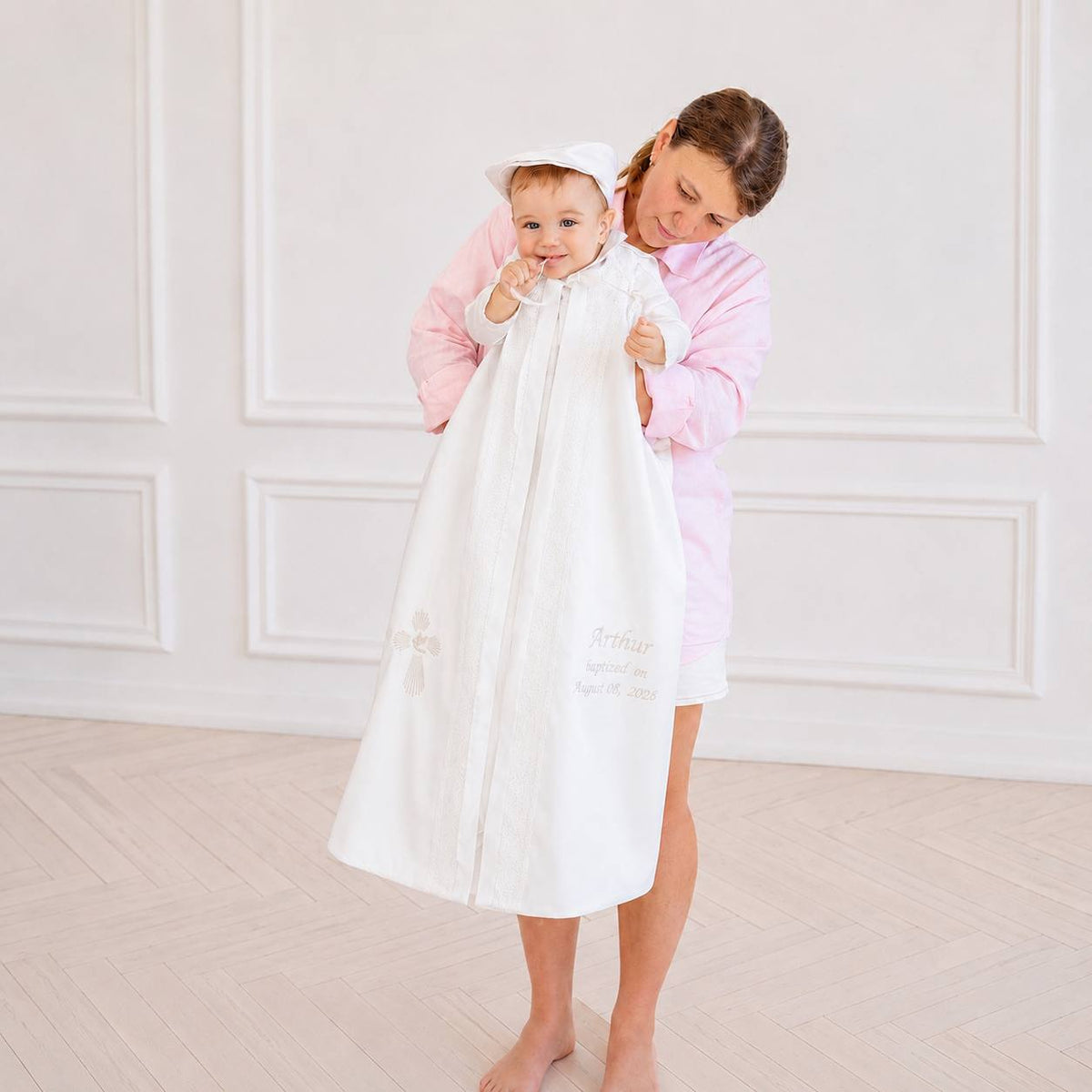A woman in a pink robe holds a baby wearing a white baptism robe with a small white hat. The room has a light wood paneled wall and pale wood floor.