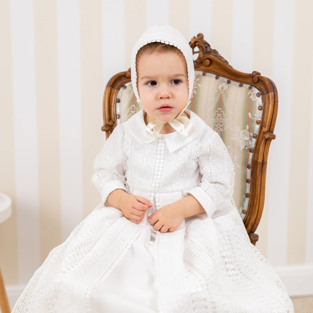 Boy in a christening gown and bonnet sitting on a wooden chair