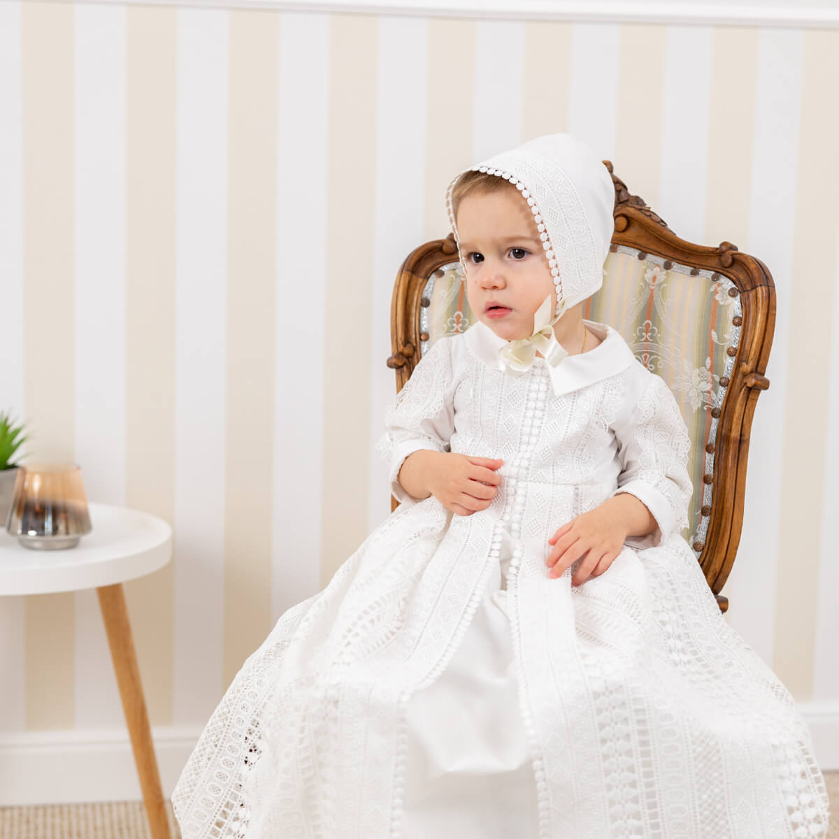 Child in a long. christening gown dress and bonnet sitting on a wooden chair