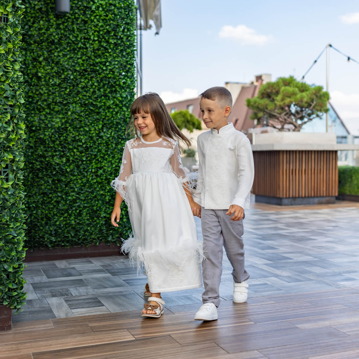 Two children in formal attire walking on a wooden deck with greenery in the background