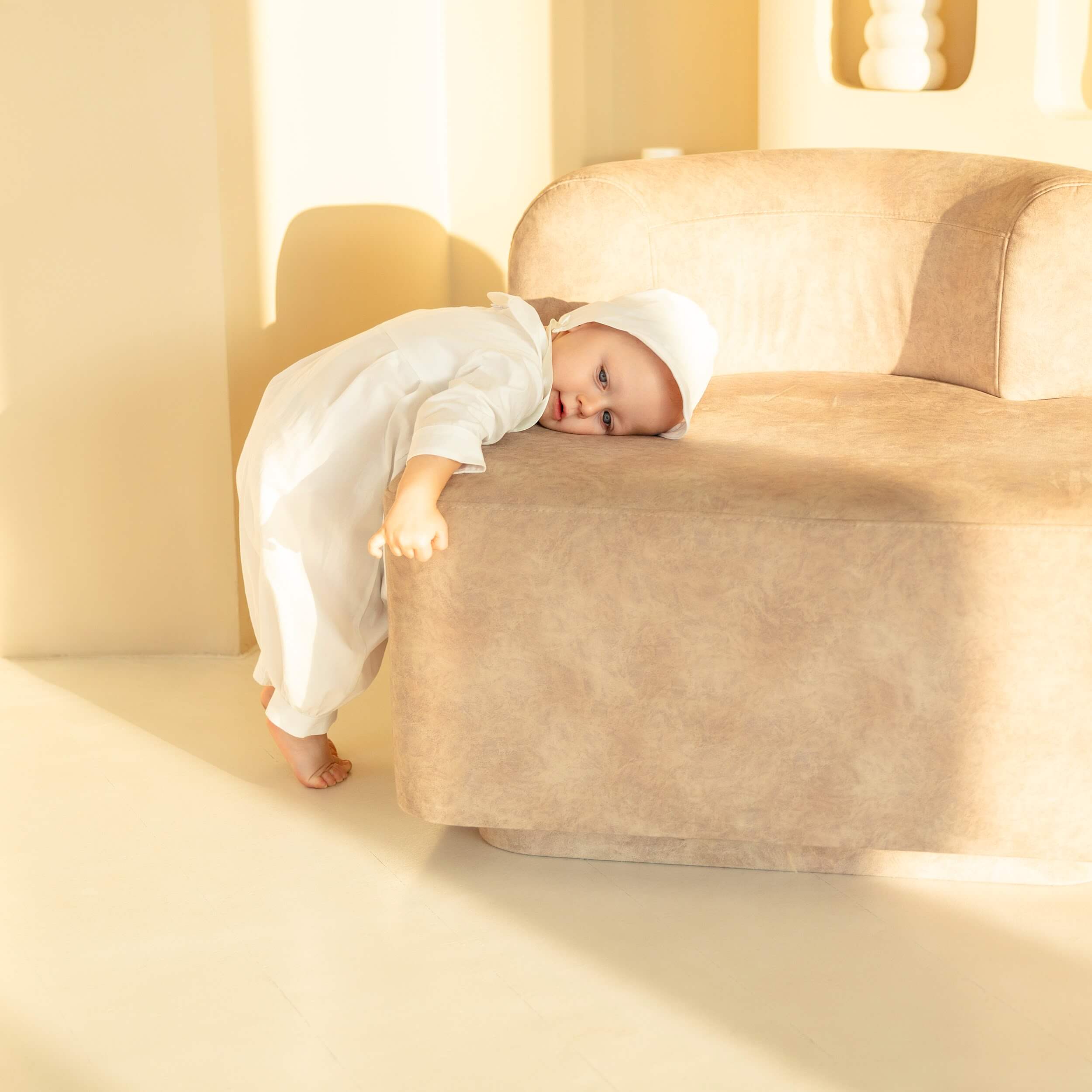 Baby in a Baby Boy Baptism Bubble Romper lying on a beige sofa in a softly lit room.