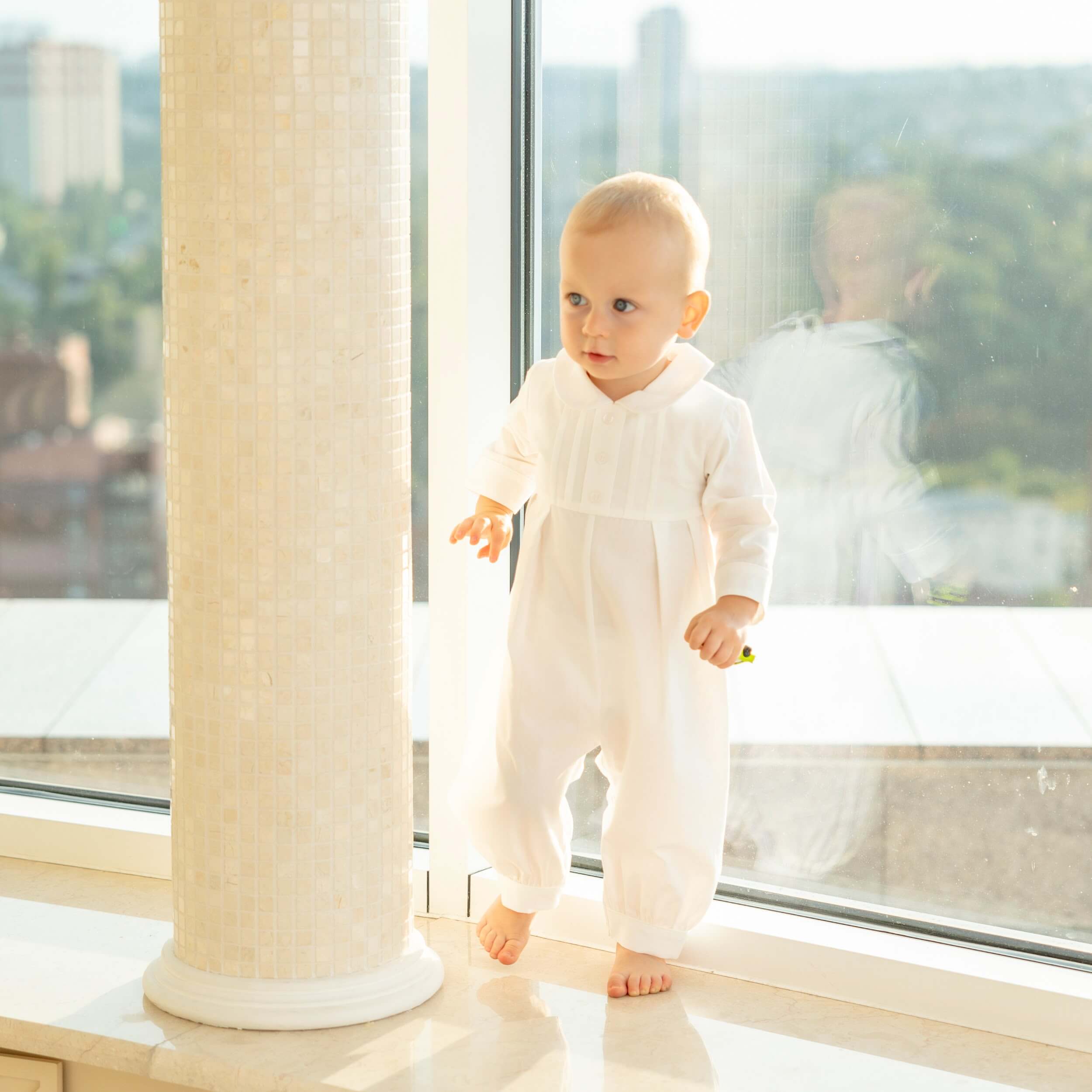 Baby in a Baby Boy Baptism Bubble Romper standing in a bright room with large windows.
