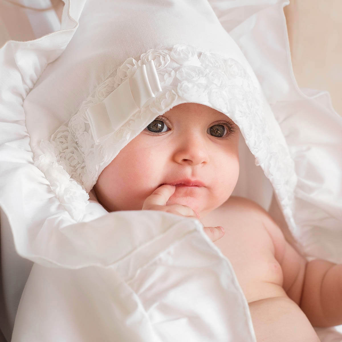 Baby wrapped in white christening blanket with a lace bonnet, looking up.