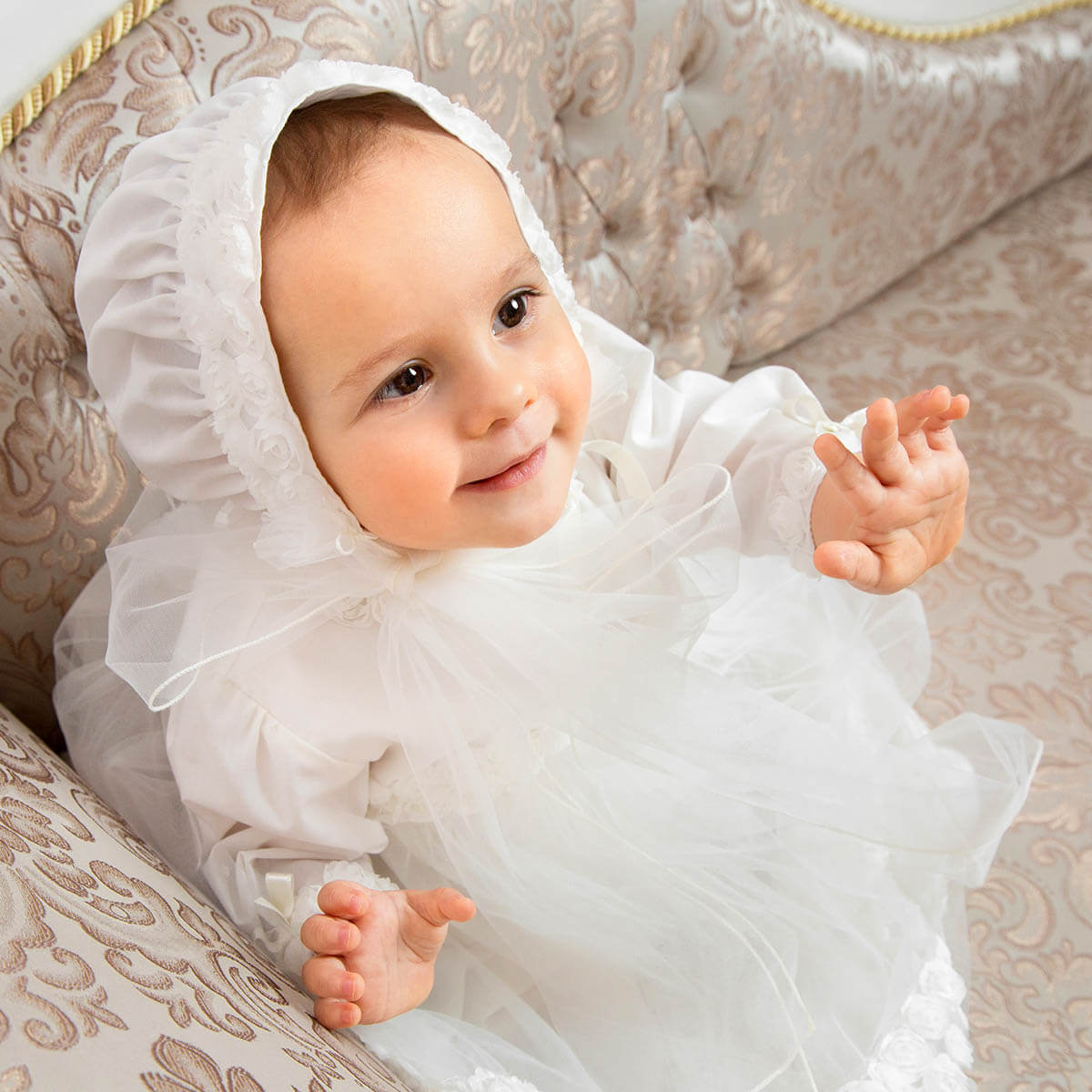Baby in a white outfit with aVintage Baptism Bonnet sitting on an ornate chair.