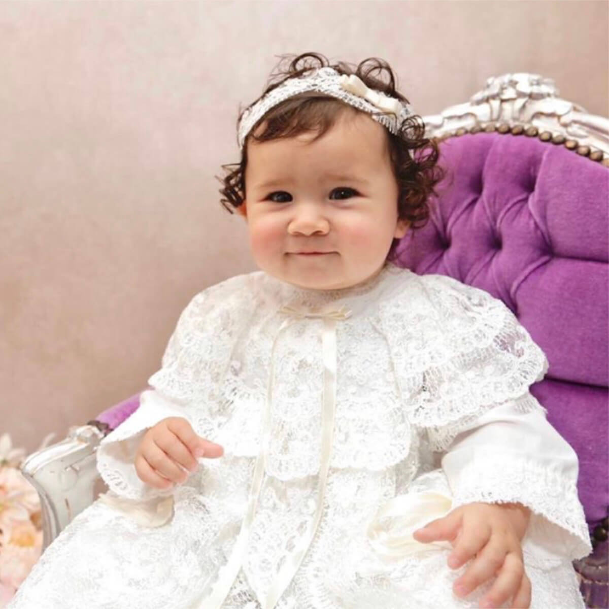 Baby in ivory christening lace outfit sitting on a purple cushion