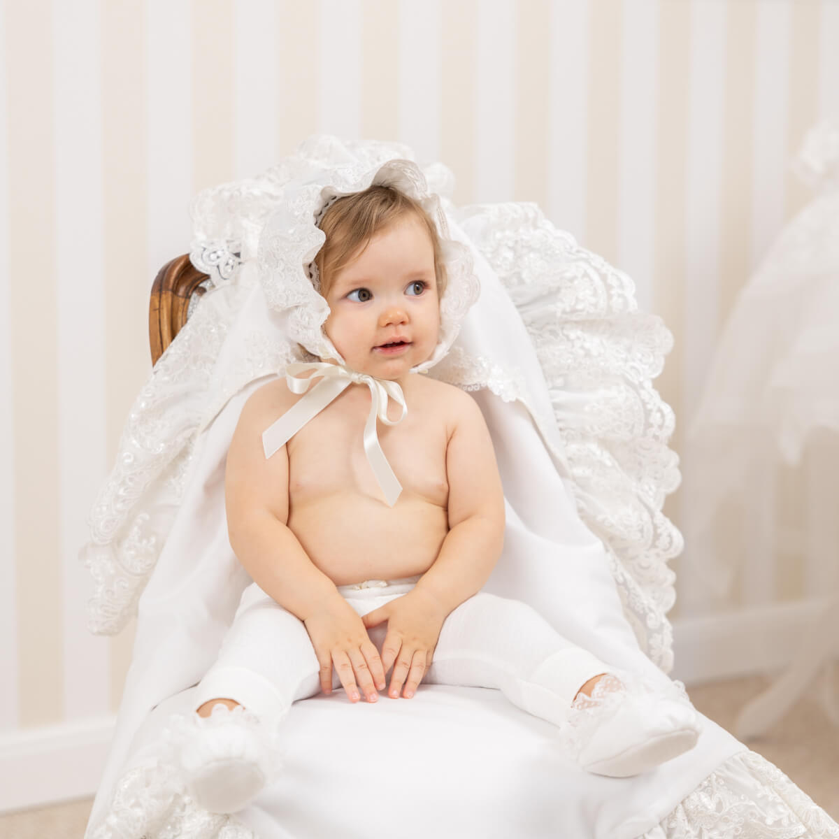 Baby wearing a white lace bonnet and dress sitting on a white chair.