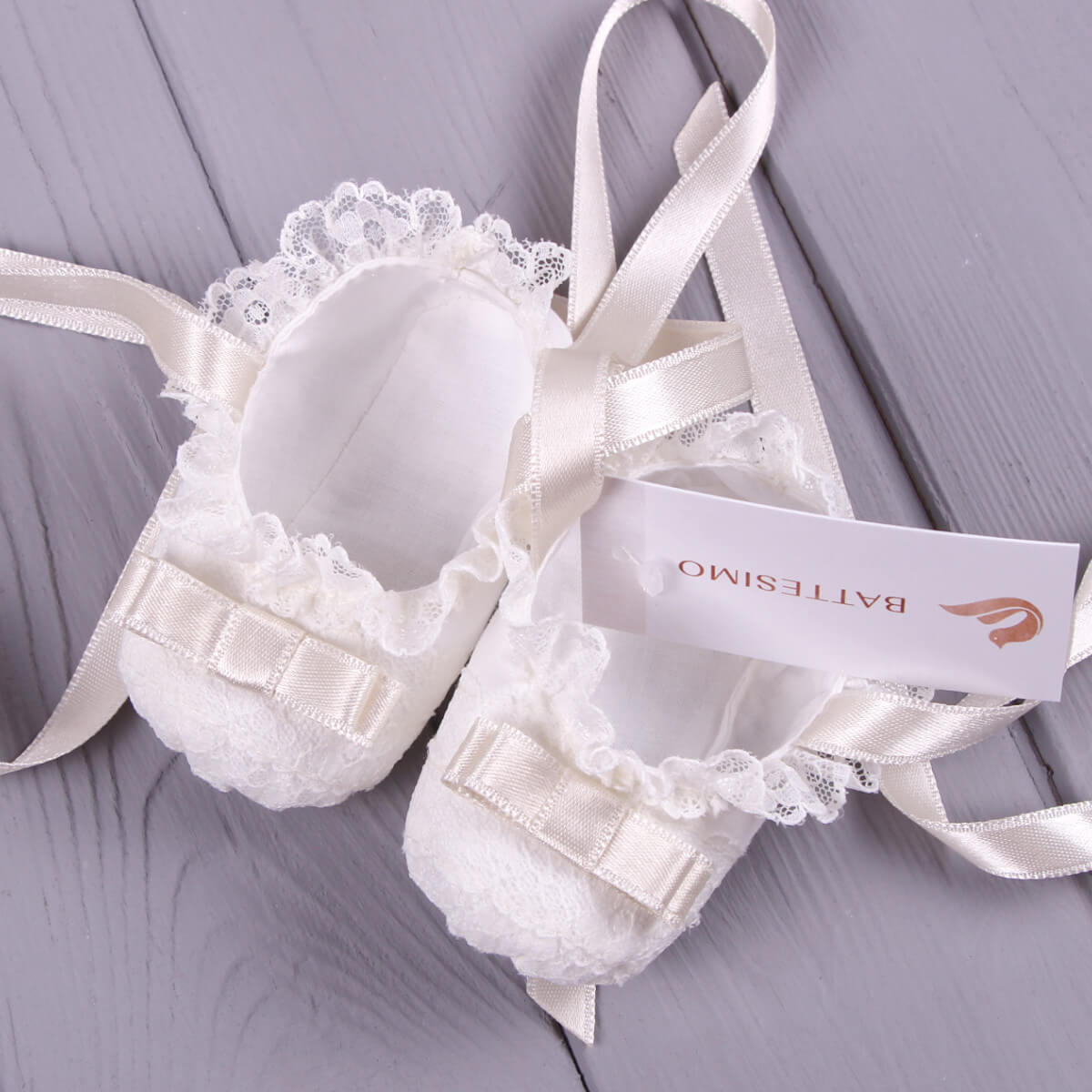 Christening Booties with ribbons and lace on a gray wooden surface, featuring a brand tag.