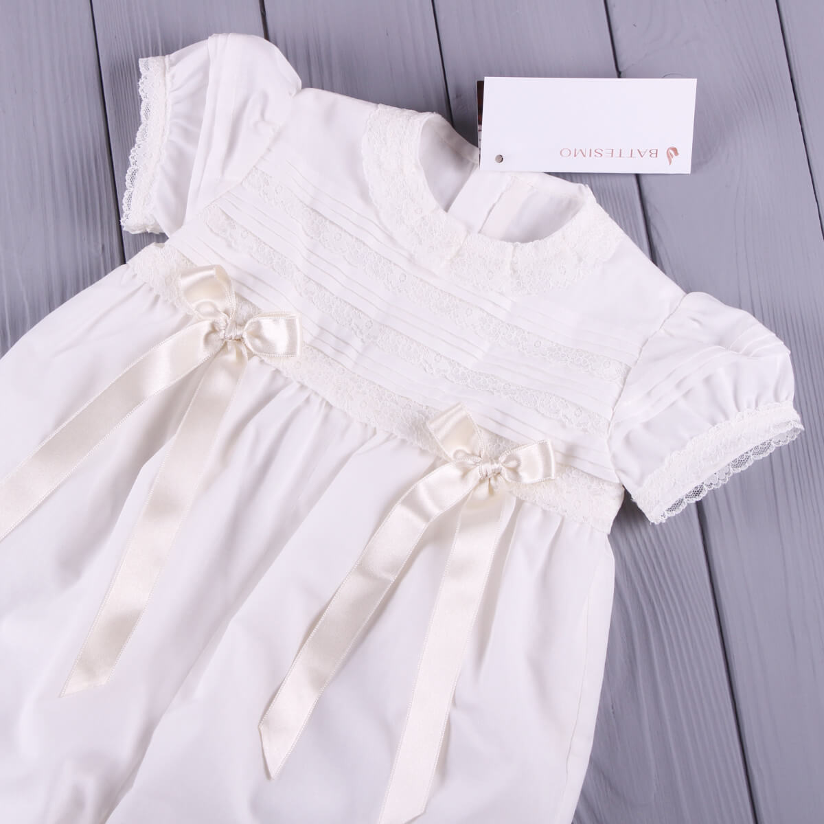 Christening romper with ribbons on a gray wooden surface, featuring the brand 'Olives Luve'.