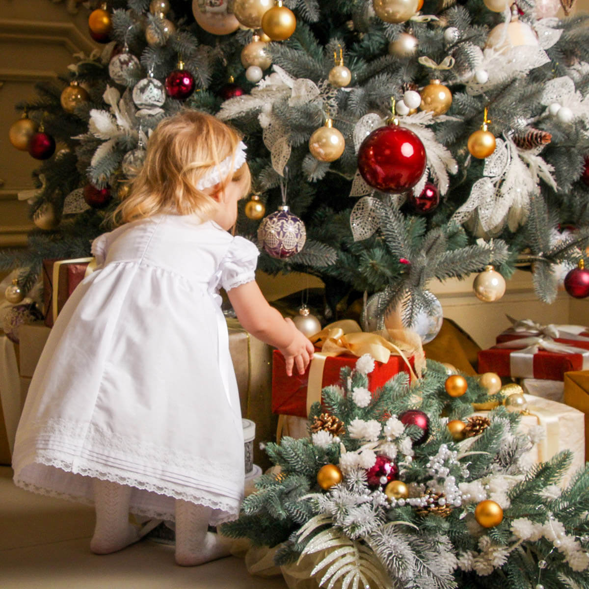Child in a Cotton Christening Dress standing next to a decorated Christmas tree with ornaments and presents.