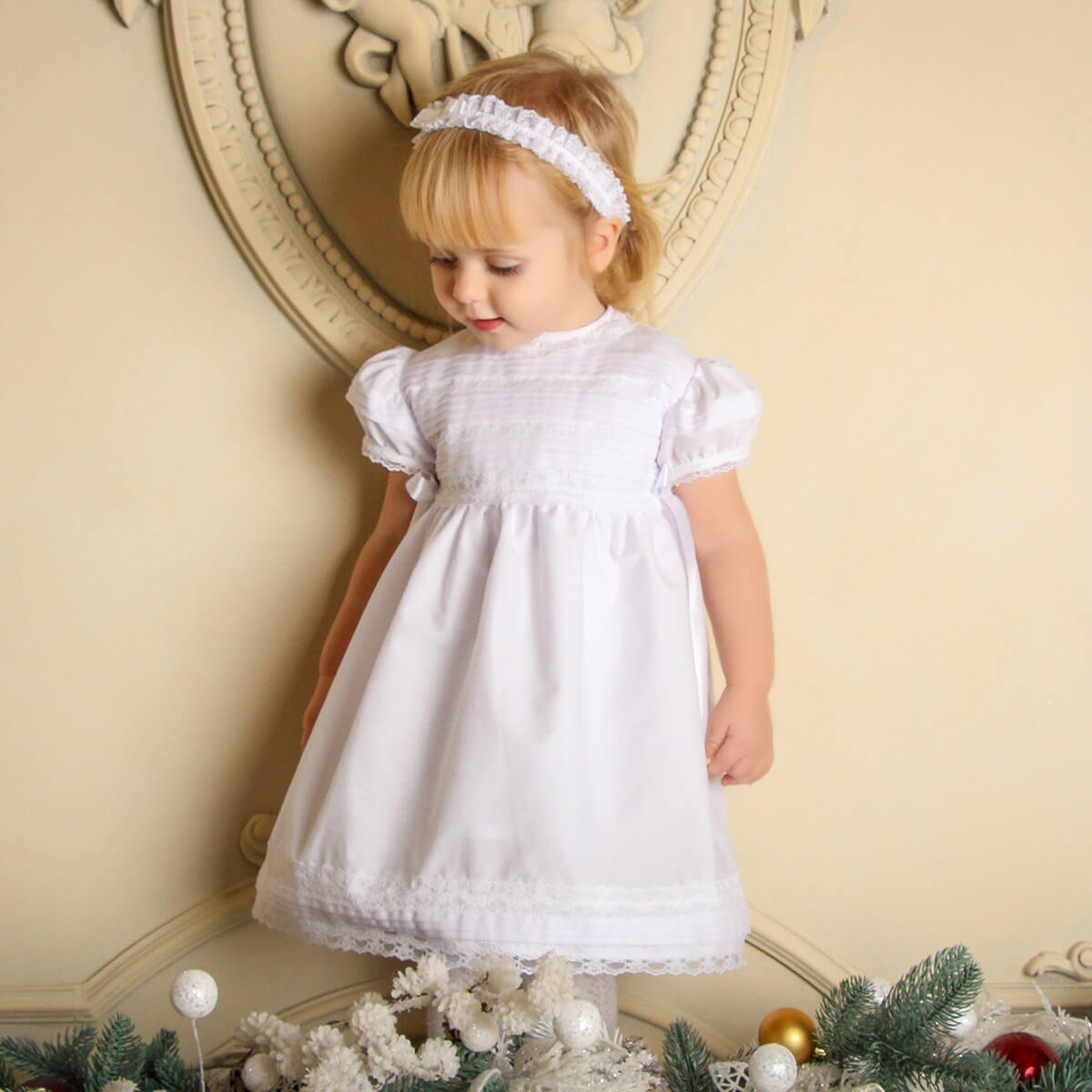 Young girl in a Cotton Christening Dress standing in front of a decorative wall with Christmas decorations.