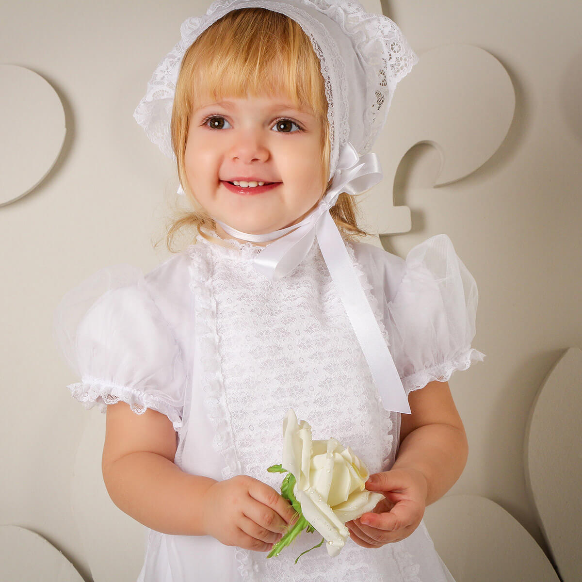 Child in a white dress with a Baby Baptism Bonnet holding a white flower against a neutral background