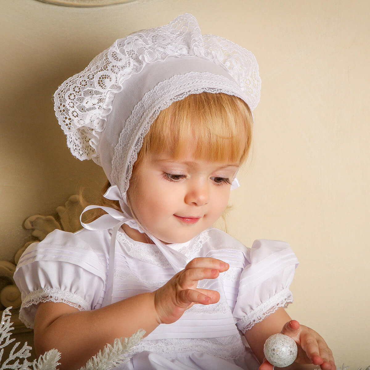 Child wearing a Baby Baptism Bonnet and dress, holding a decorative item.