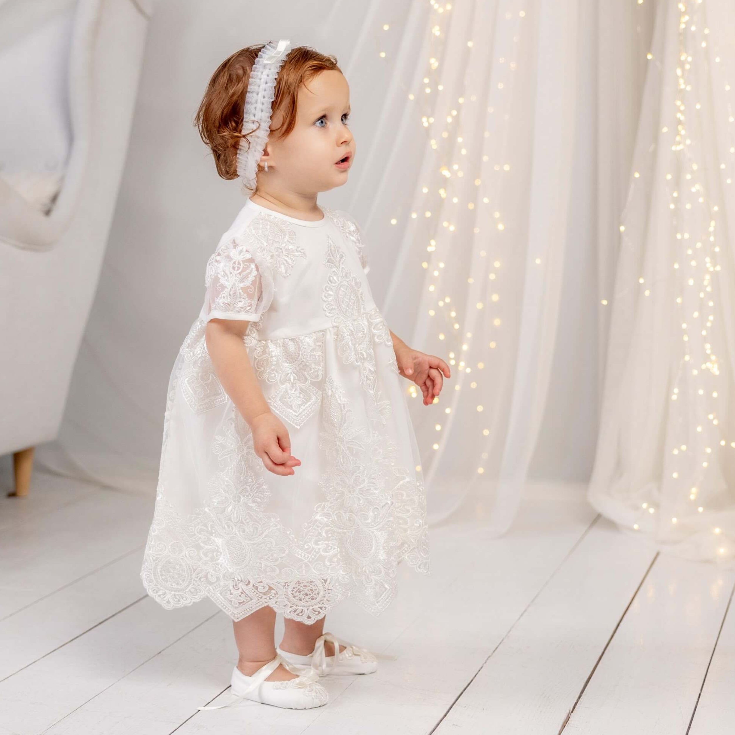 Young child in a white lace dress standing in a softly lit room with white curtains and furniture.