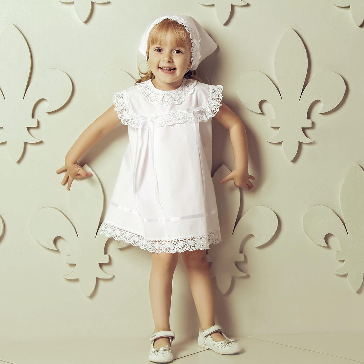 Young girl in a Summer Baptism Dress Set for Girl with lace details standing against a decorative wall.