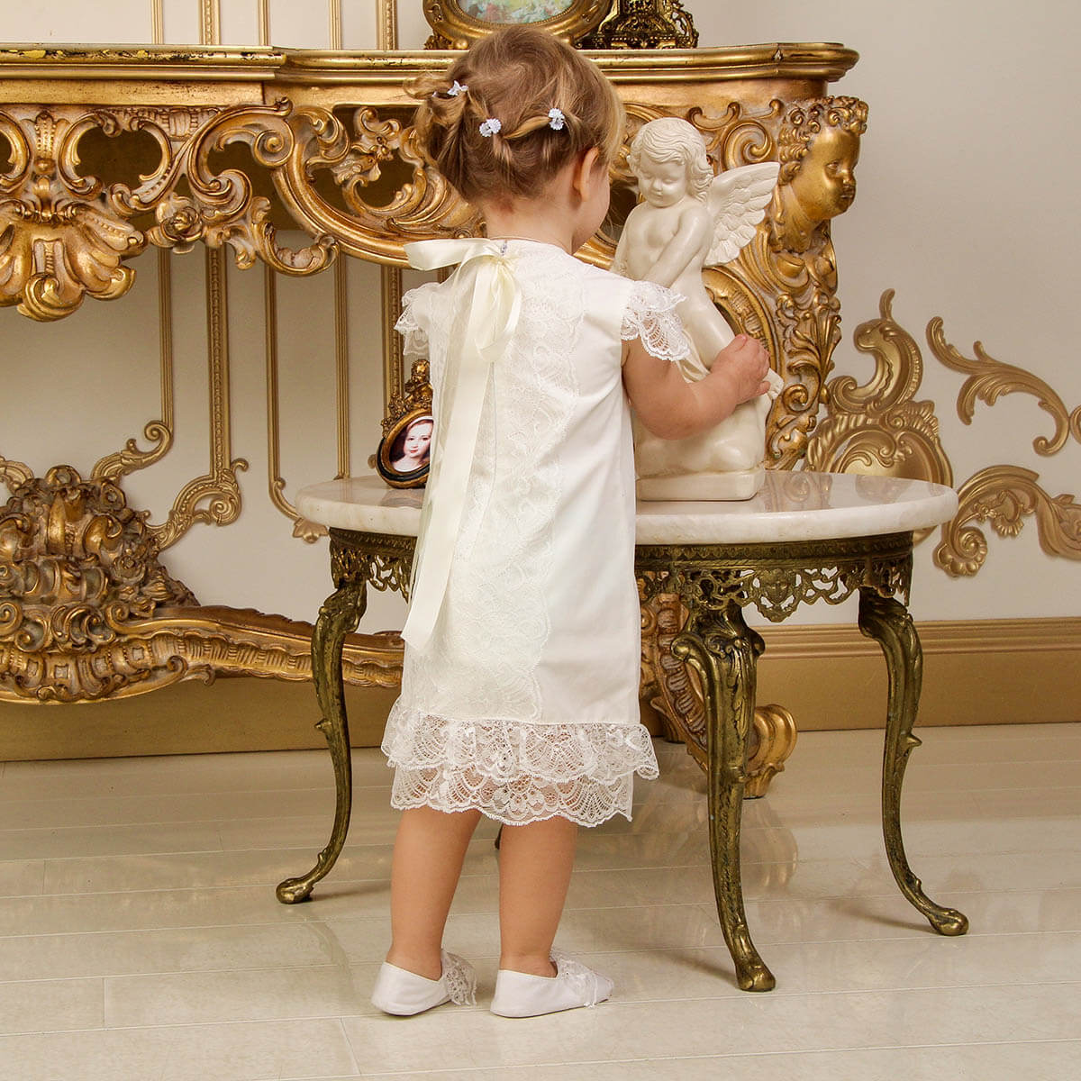 Young girl in a white dress standing next to an ornate gold table with a marble top.