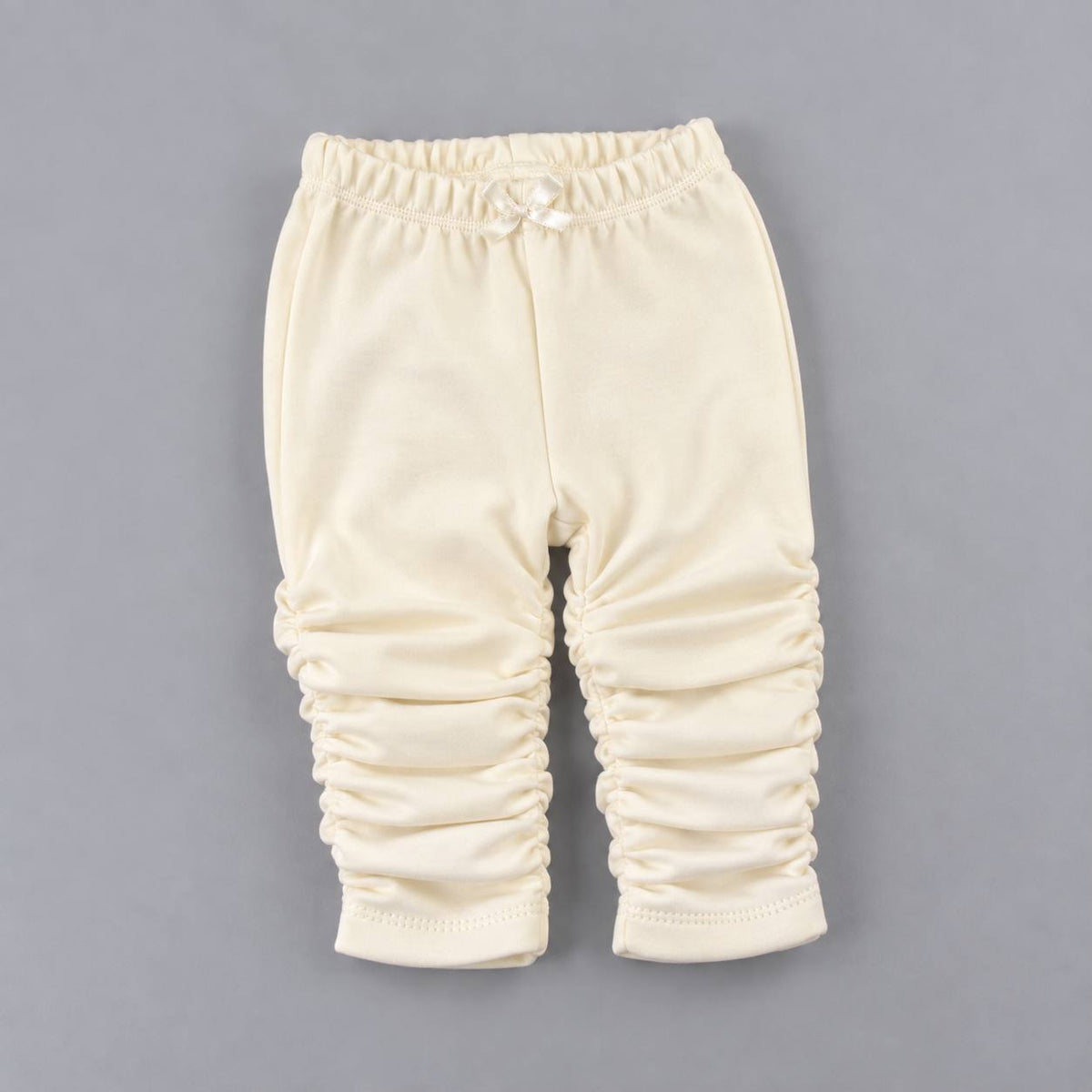 Cream-colored baby pants with ruched leg detailing and an elastic waistband, laid flat on a neutral gray background.