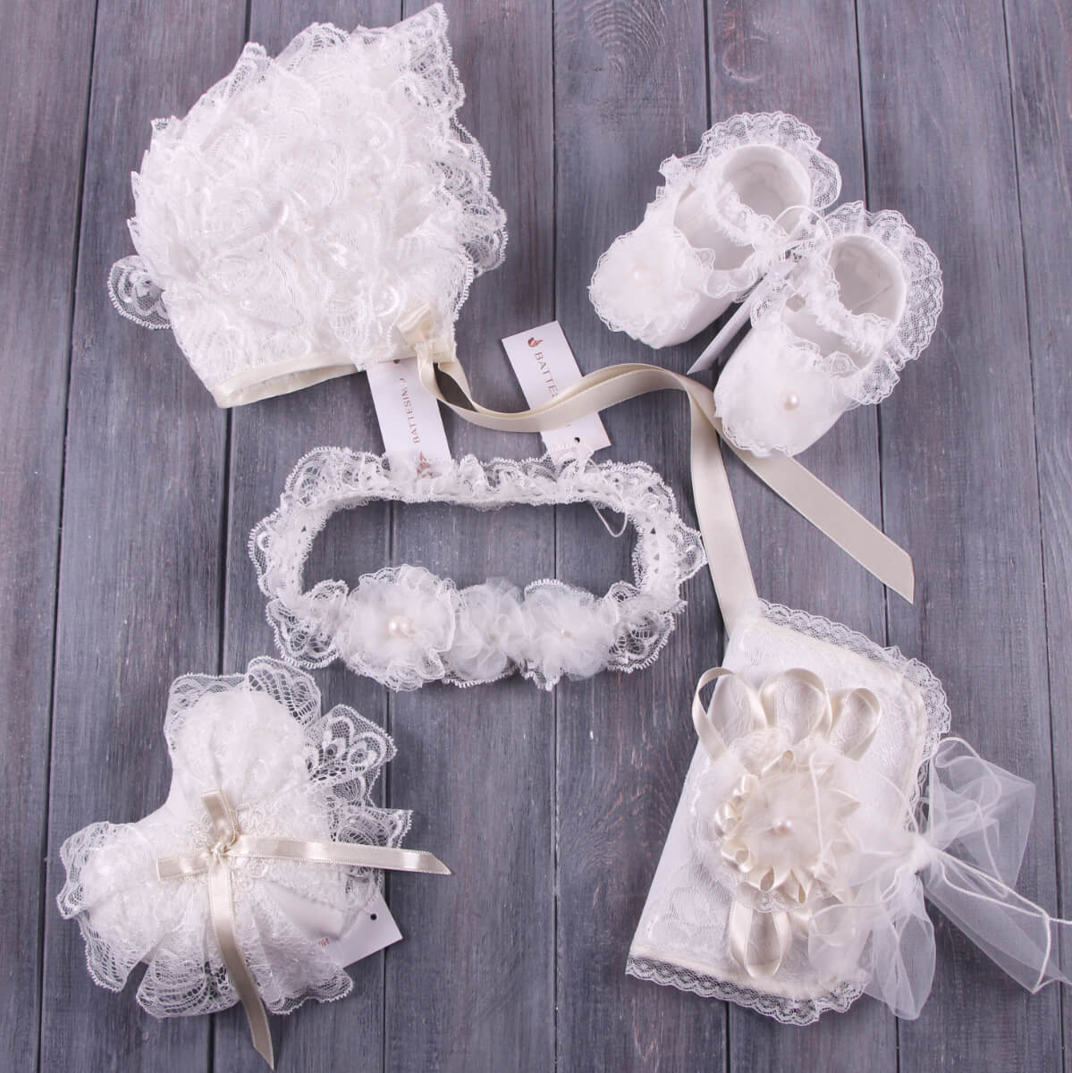 Set of white lace baby accessories including shoes, lace baptism bonnet and booties on a wooden surface.