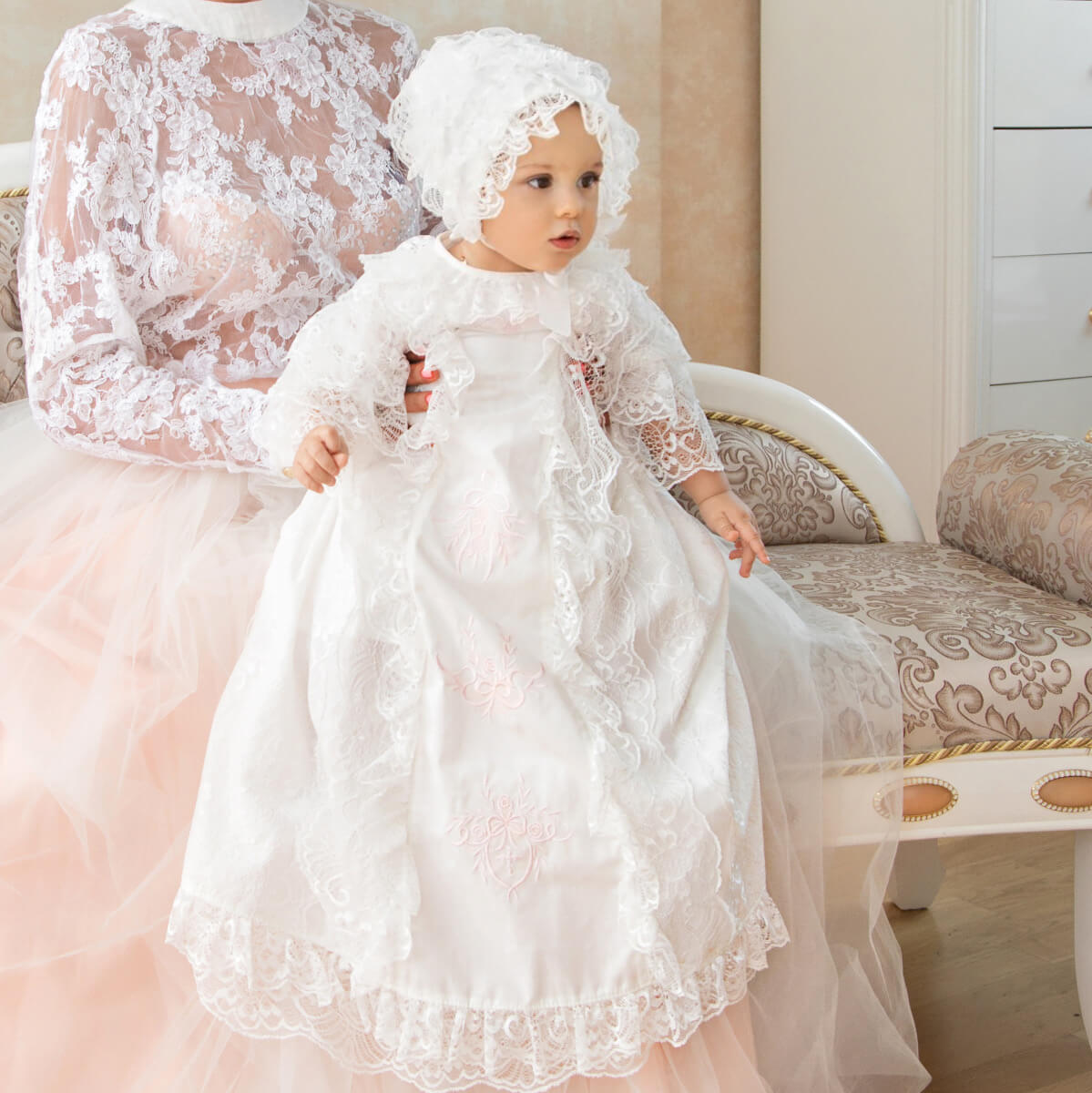 Baby in a Christening Gown for Girls sitting on a decorative chair with a blurred figure in the background.