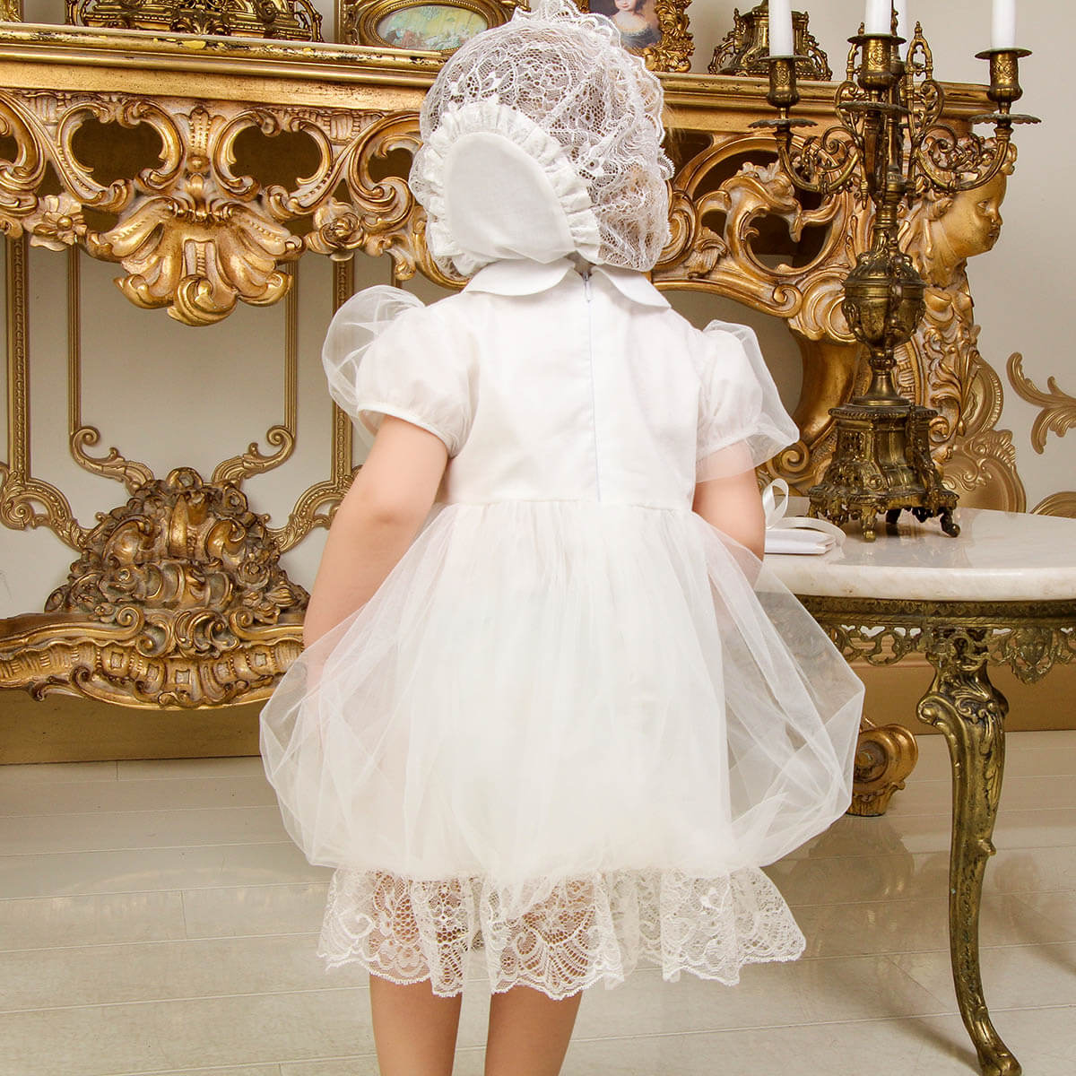 Child in a white lace dress and Lace Baptism Bonnet standing in an ornate room.