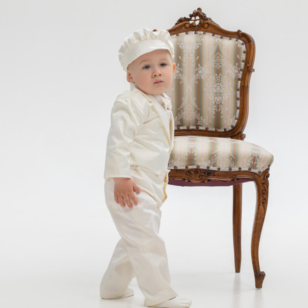 Baby in a Baby Boy Baptism Suit standing next to an ornate chair on a white background