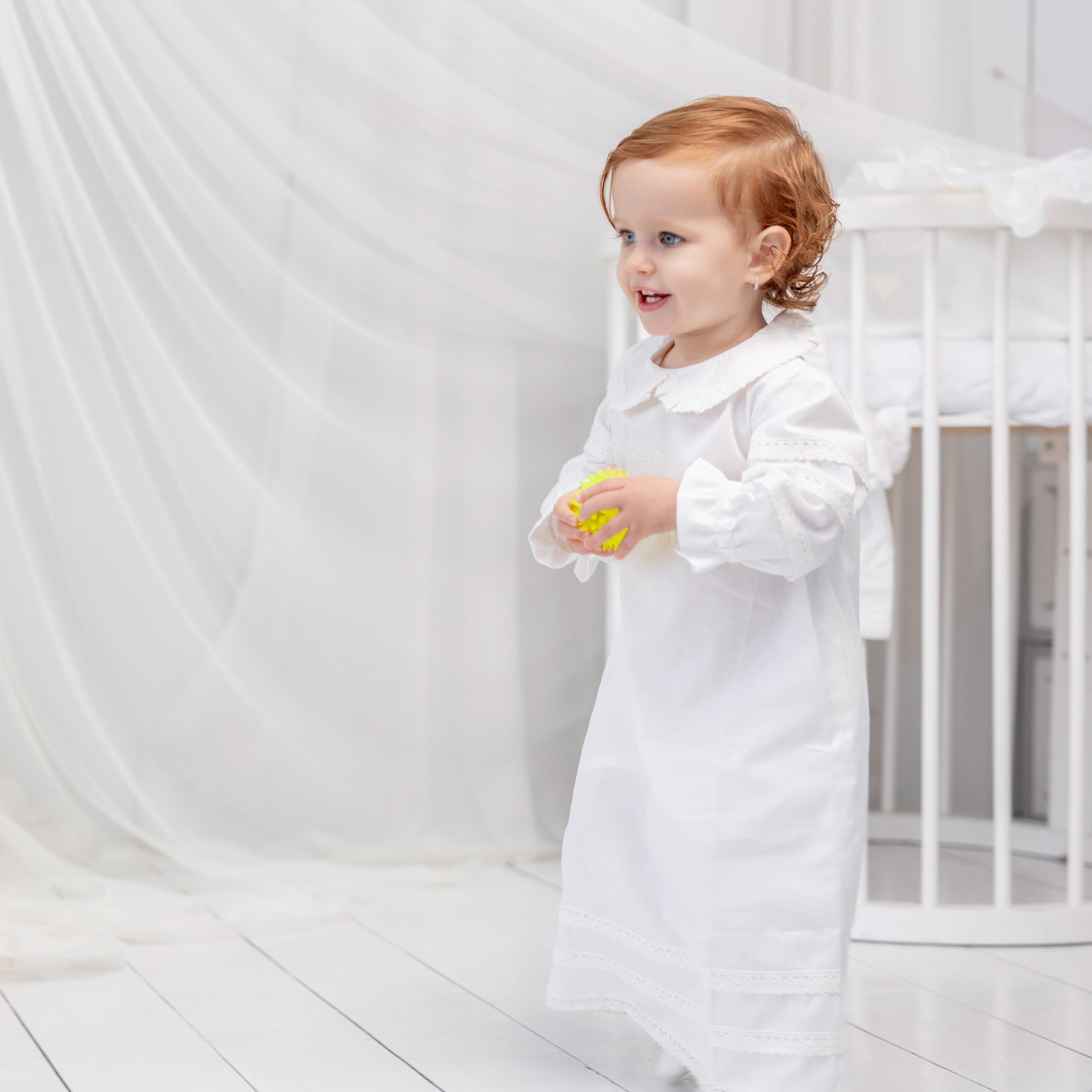 Child in a Baptism Gown standing in a minimalistic room with white curtains.