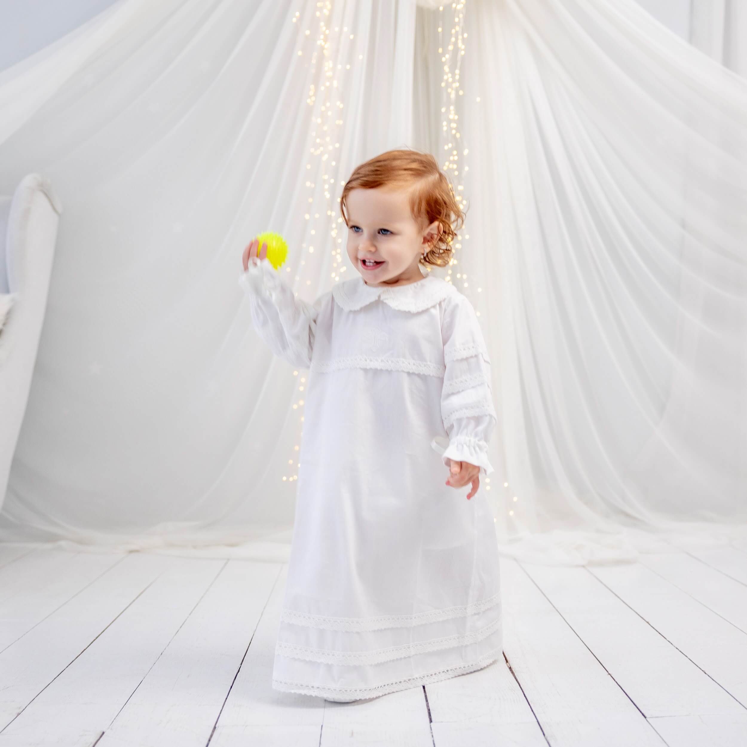 Child in a Baptism Gown holding a yellow ball against a white background with string lights.