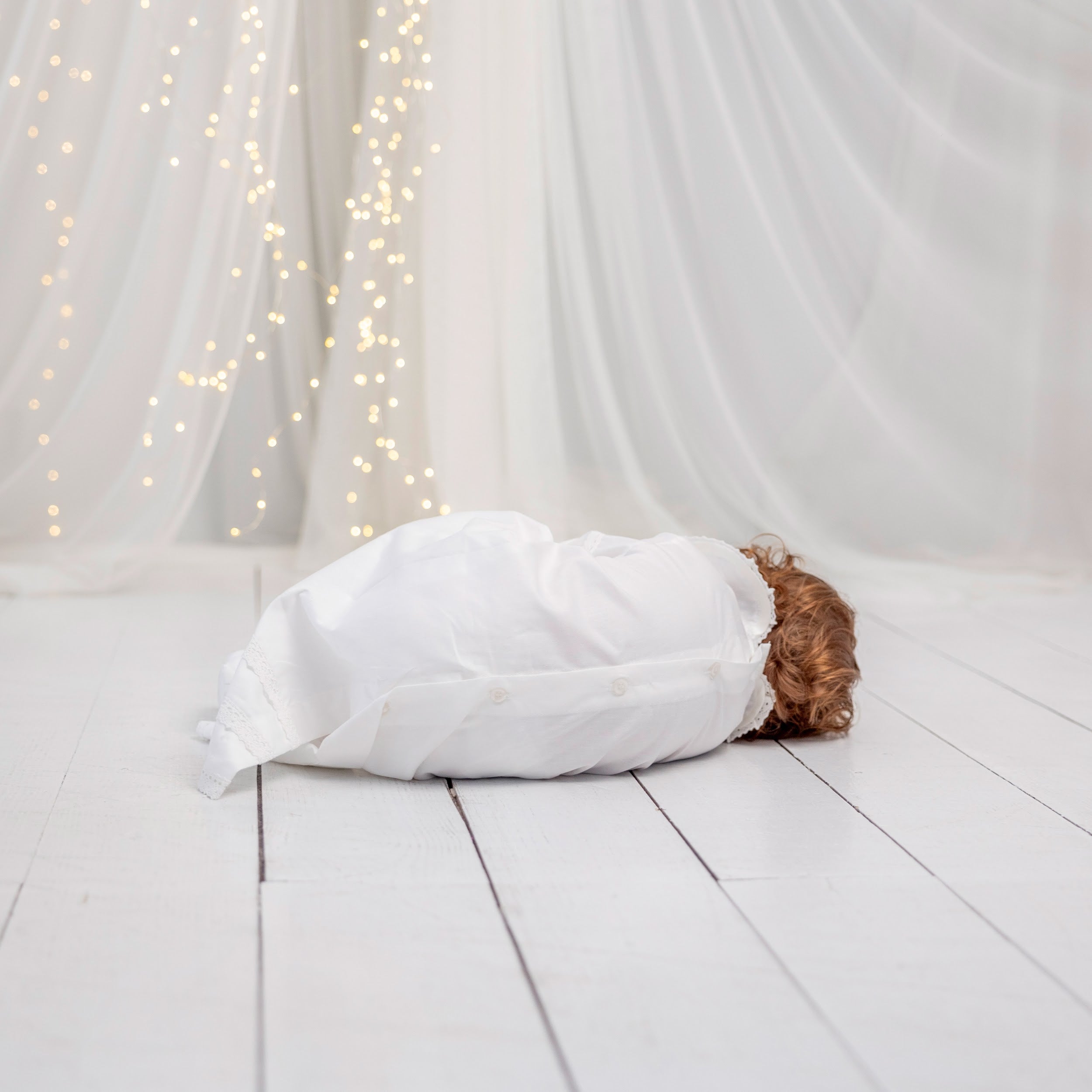 Person dressed in a Baptism Gown on a light wooden floor with white curtains and string lights in the background