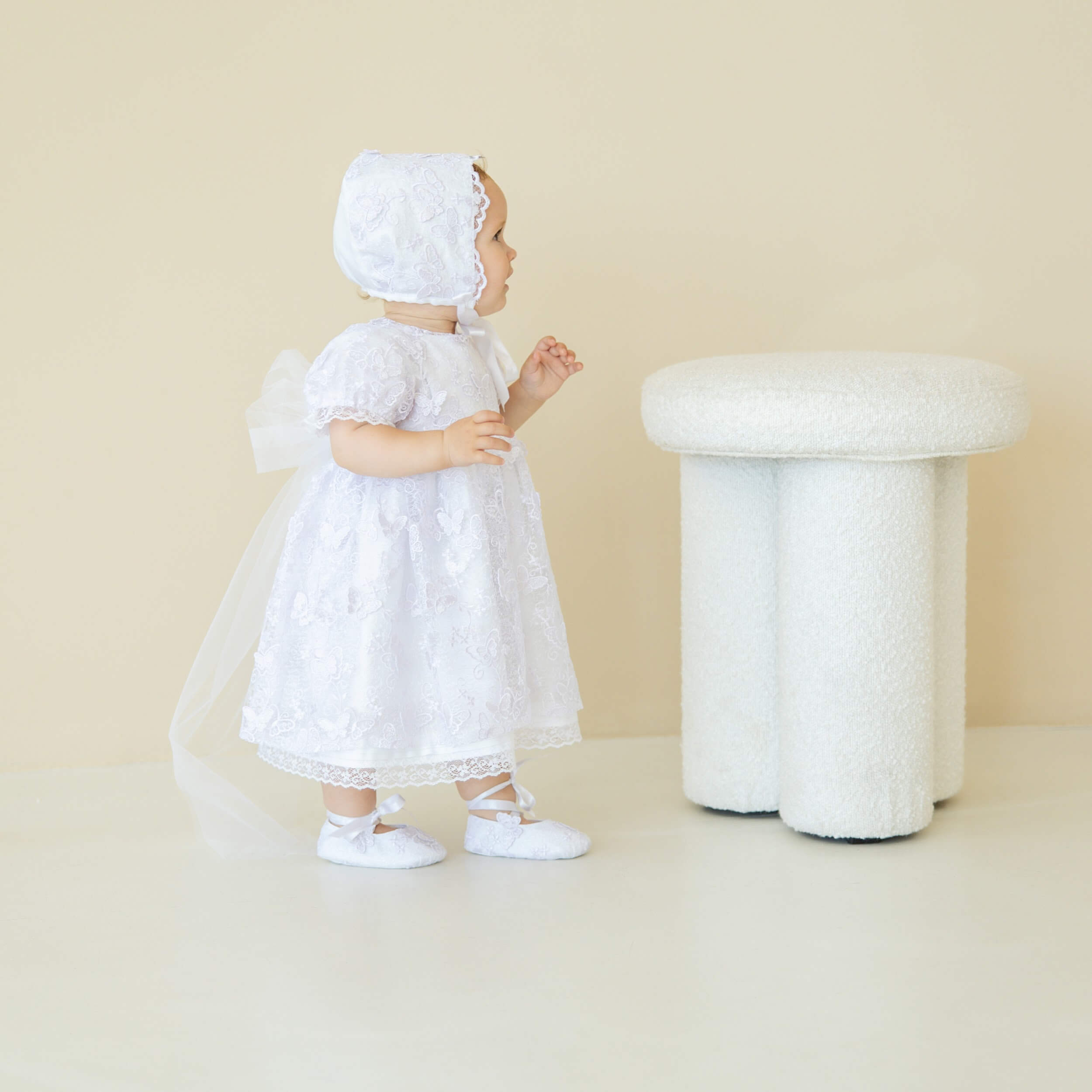 Baby in a Baptism Dress with Lace and bonnet standing next to a white mushroom-shaped stool on a beige background