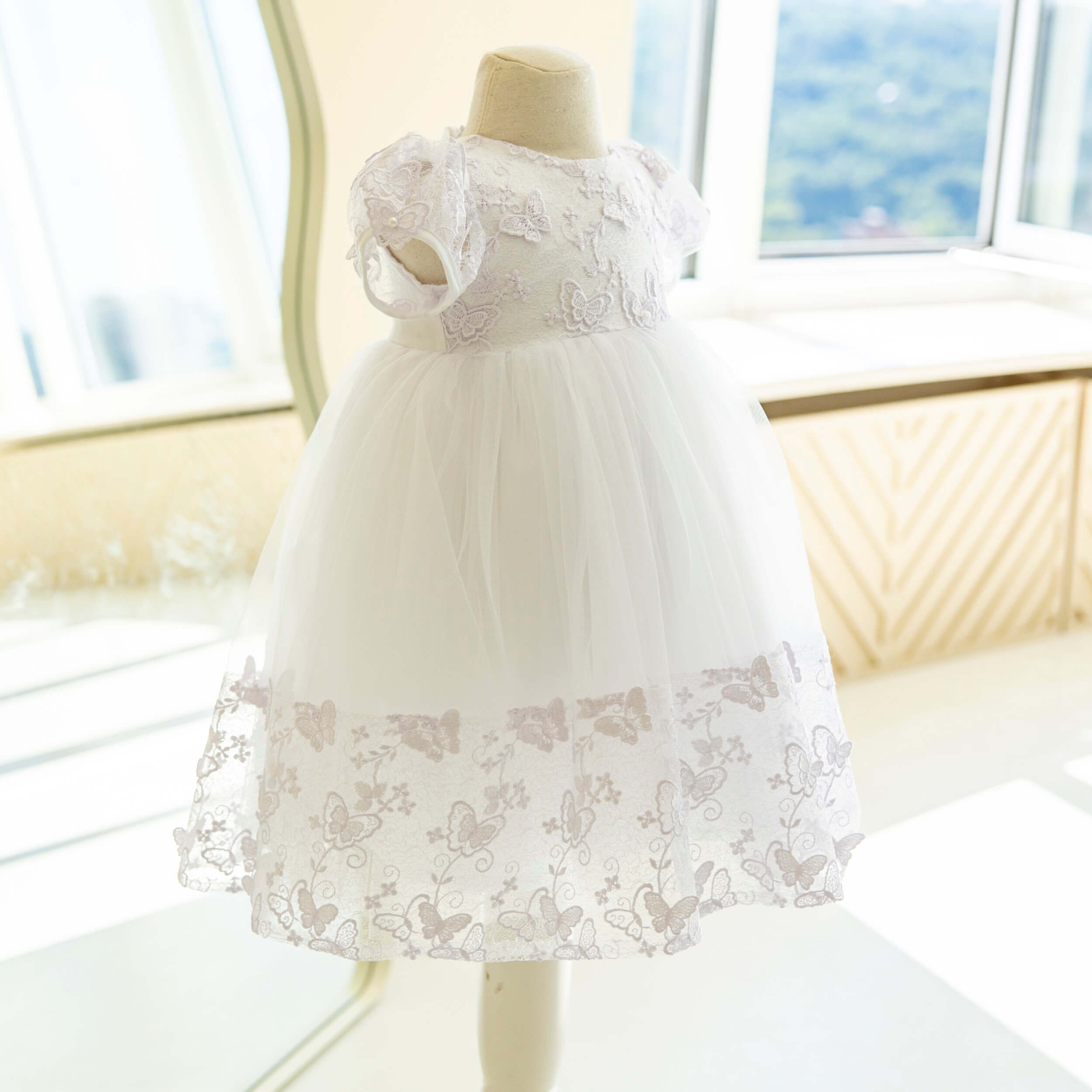 Baptism Dress with Butterfly Lace on a mannequin in a bright room with large windows.