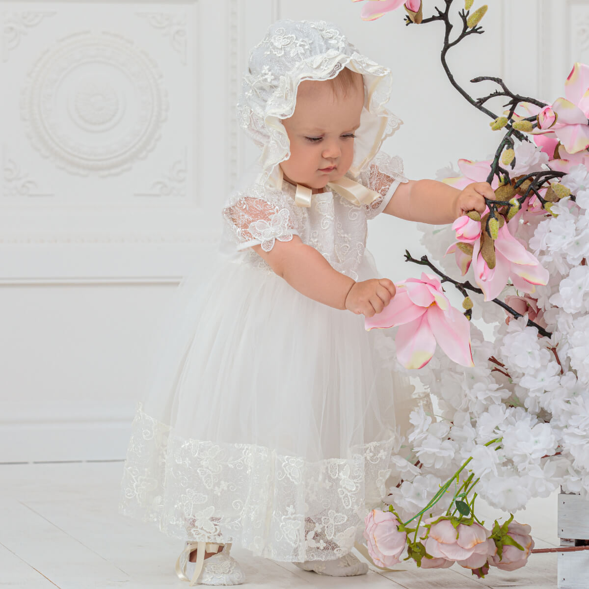 Baby in a Baptism Dress with Butterfly Lace interacting with pink flowers against a white background