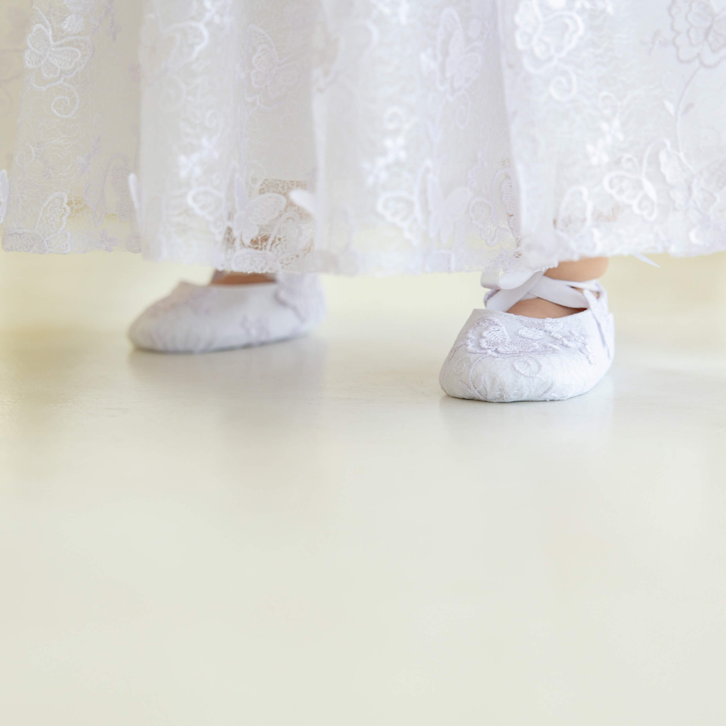 White ballet shoes on a white surface with a Baptism Dress with Butterfly Lace in the background