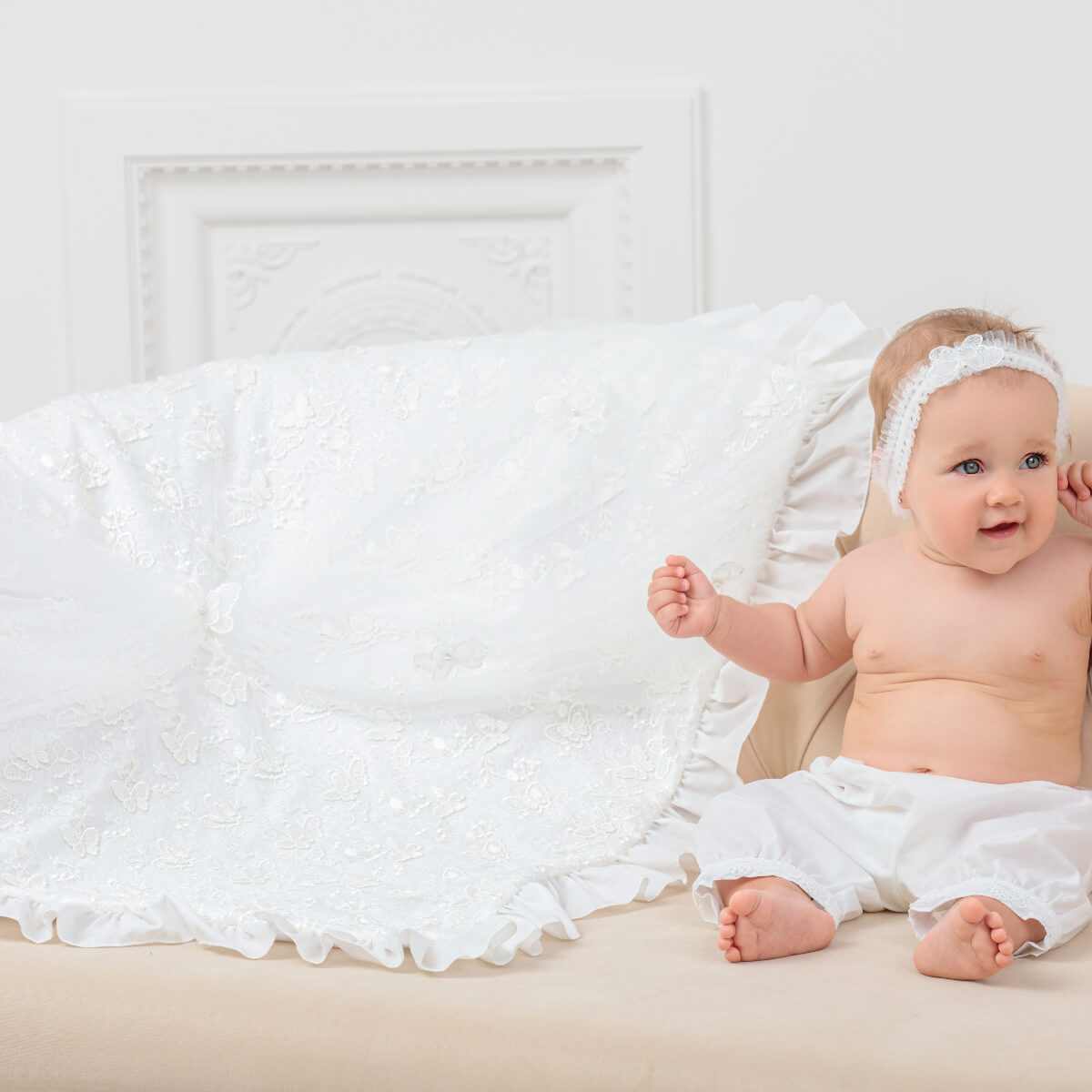 Baby sitting on a white couch with a white christening blanket and headband.