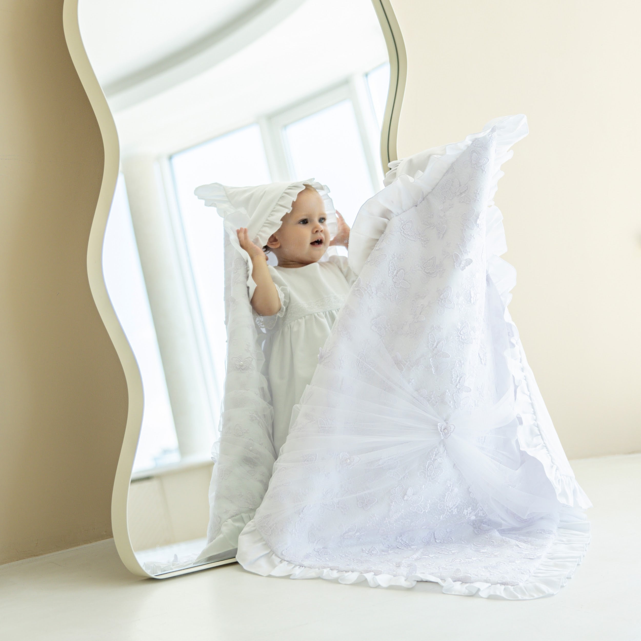 Baby playing with a large white christening blanket in a bright room.