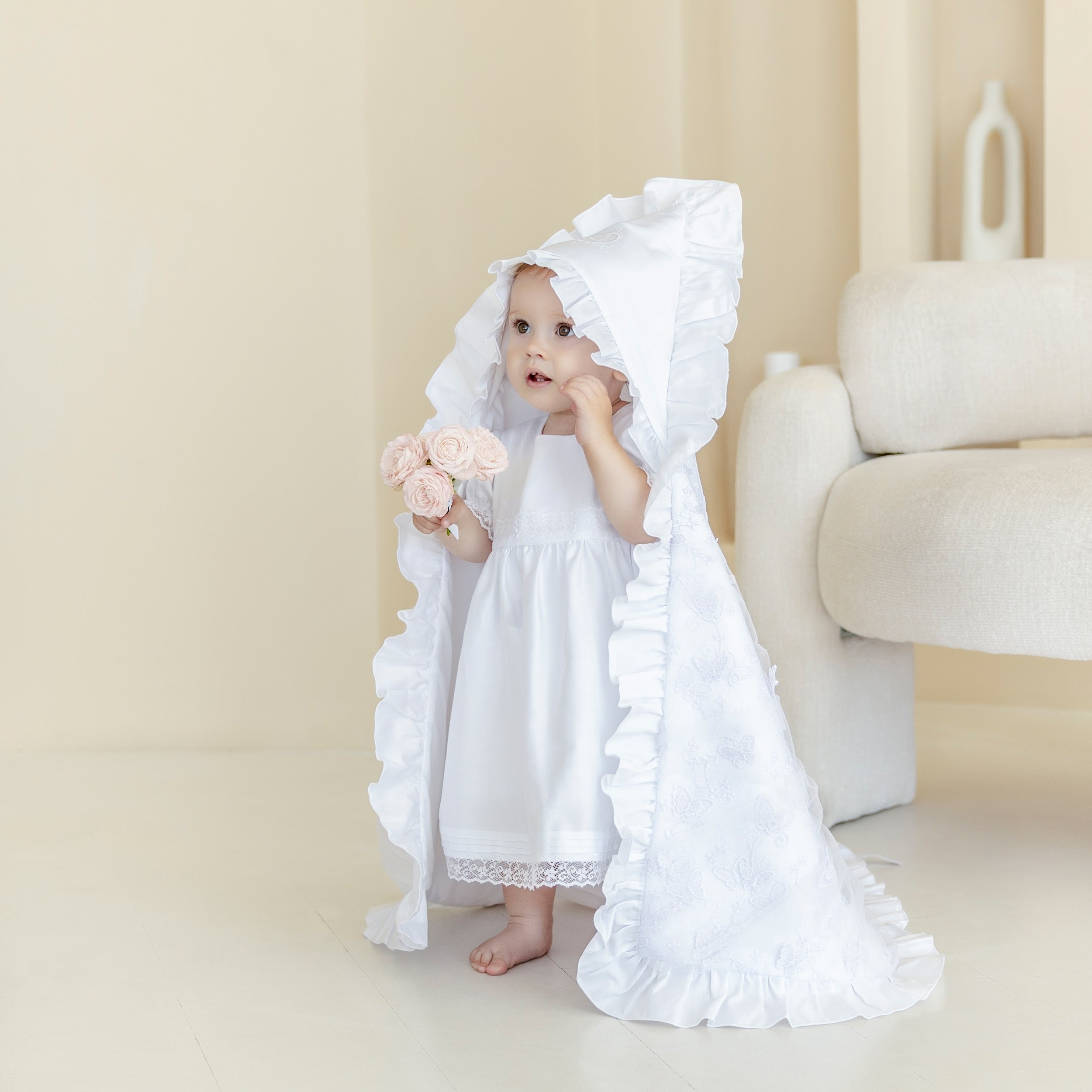 Child in a white christening blanket with a hood holding flowers indoors.