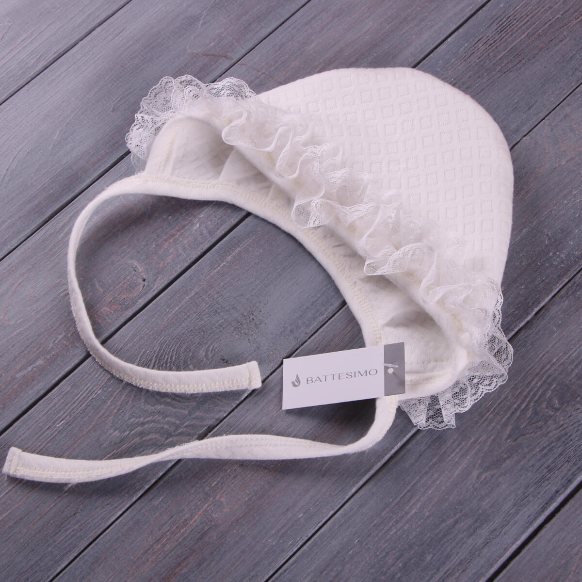 White baby bonnet with lace trim as a part of Coming Home Romper on a wooden surface, featuring a brand tag.