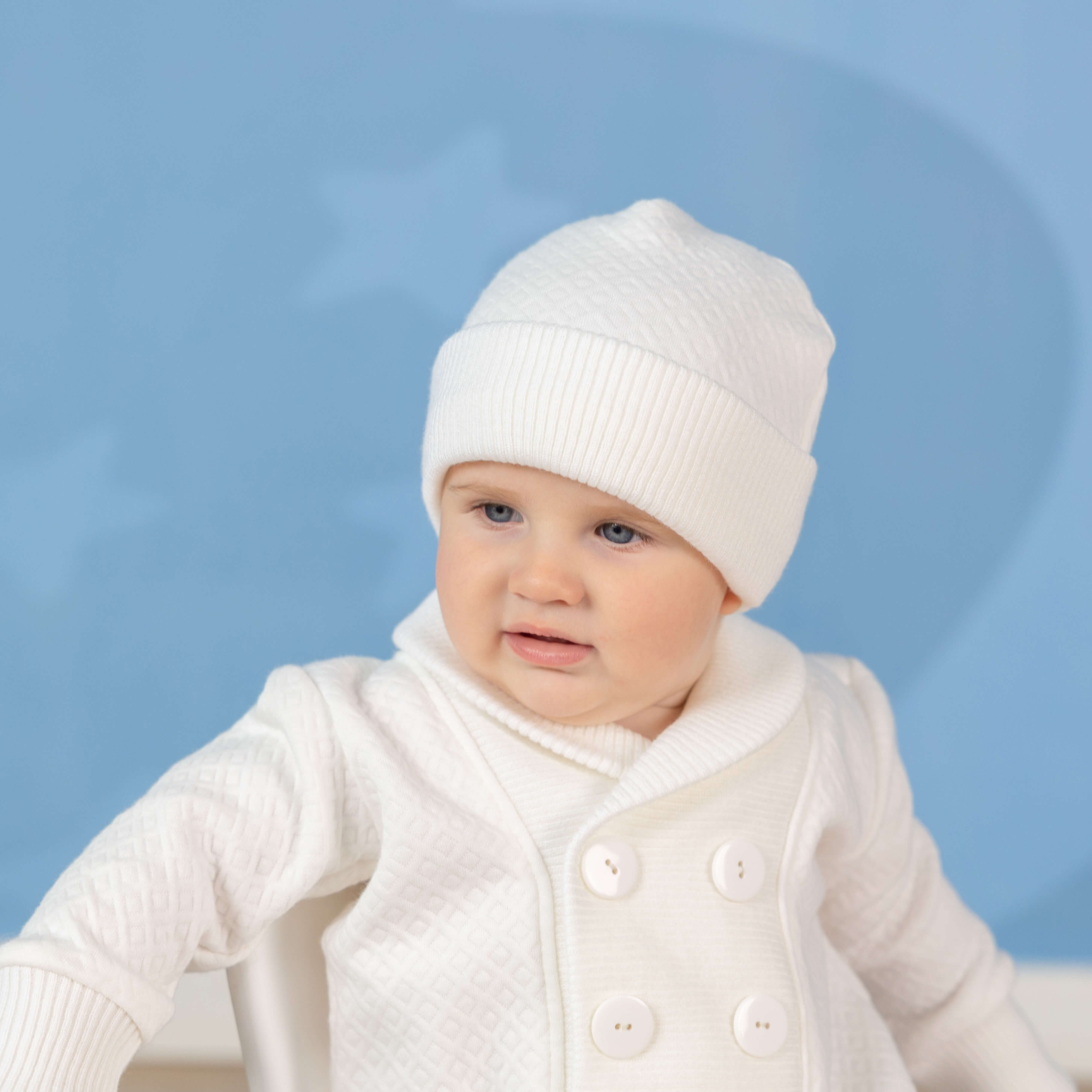 Baby wearing a white outfit and Boy Baptism Hat against a light blue background