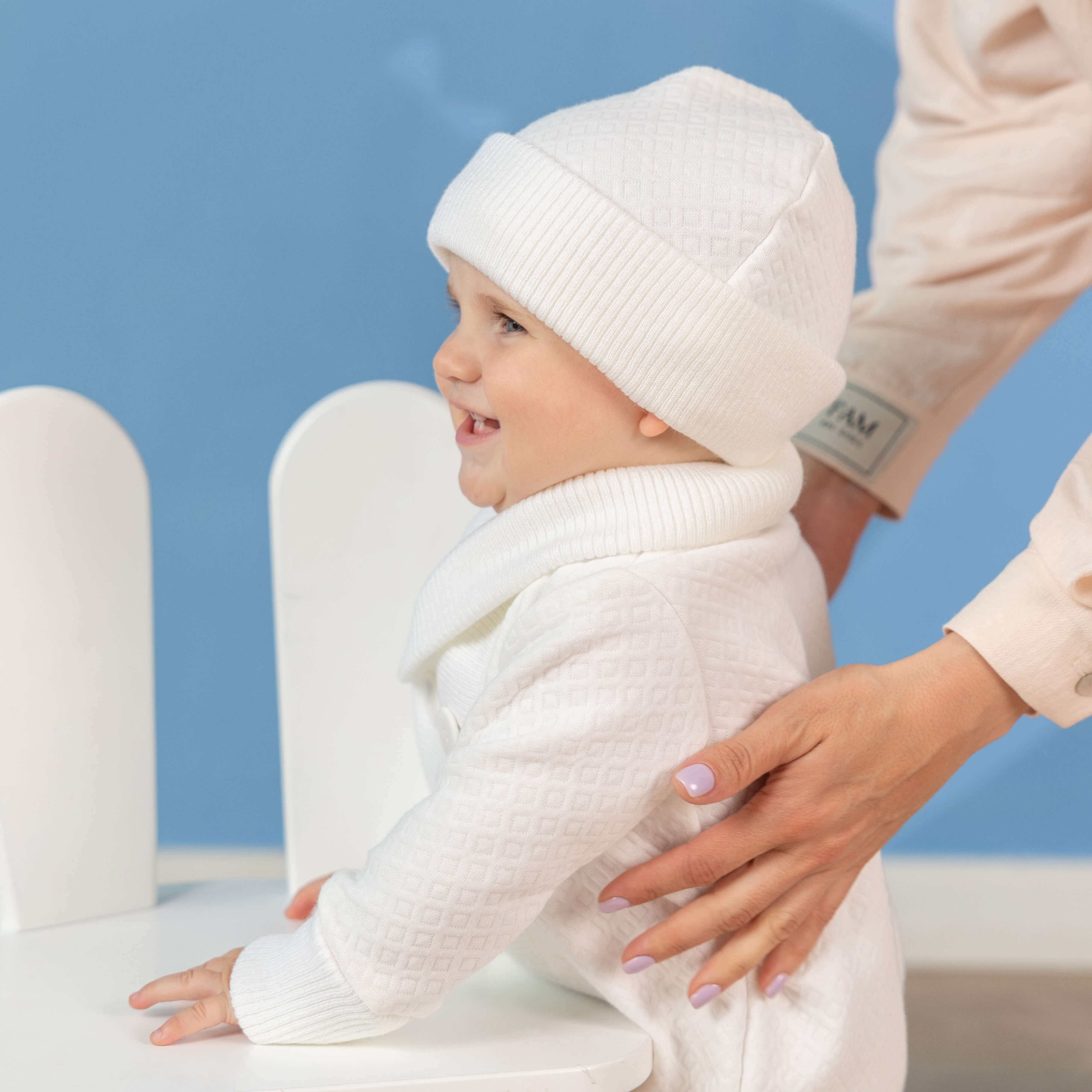 Baby in white outfit with a Boy Baptism Hat, sitting on a white chair against a blue background