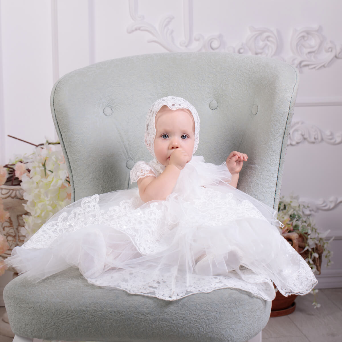 Baby in a Lace Baptism Dress with Train sitting on a gray chair with a decorative background