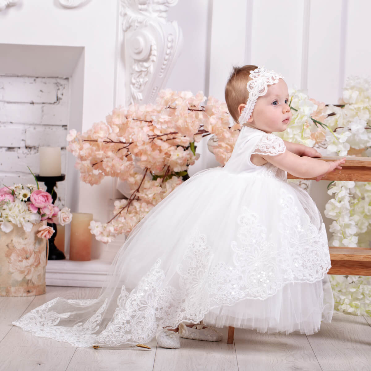 Baby in a Lace Baptism Dress with Train s sitting on a wooden stool with floral decorations in the background.