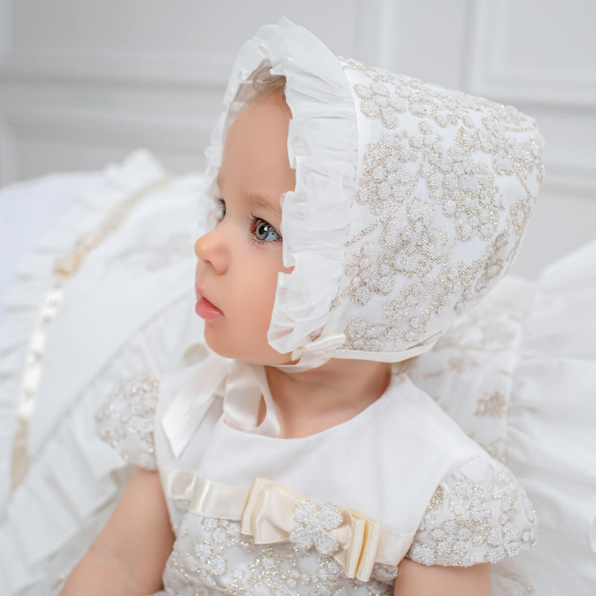 Baby wearing a Christening Bonnet and dress against a neutral background