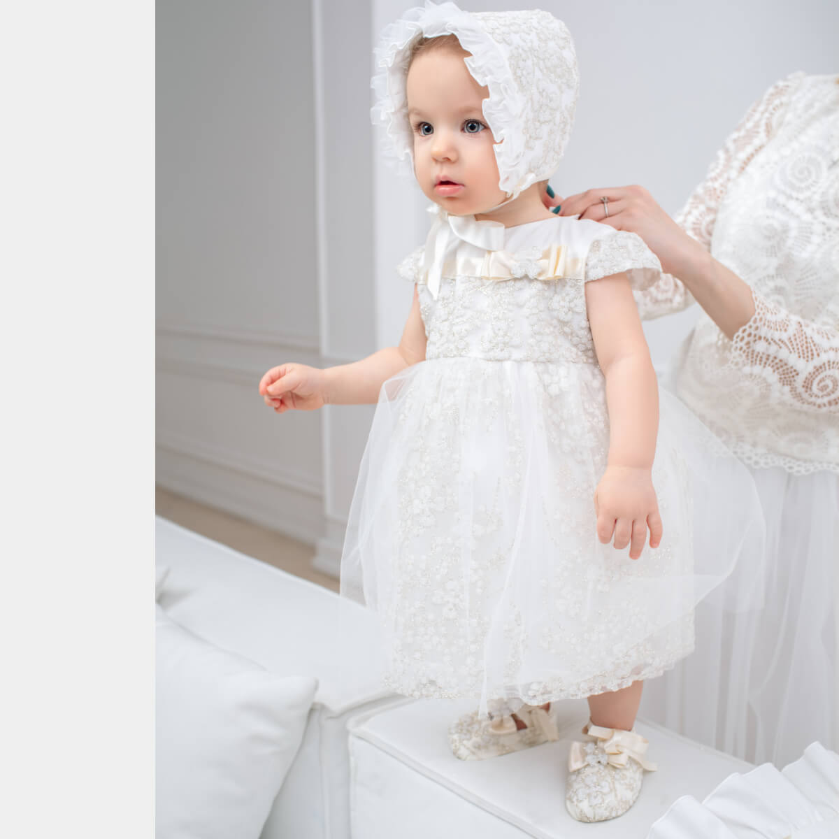 Baby in a Baby Baptism Dress and bonnet standing on a white surface.