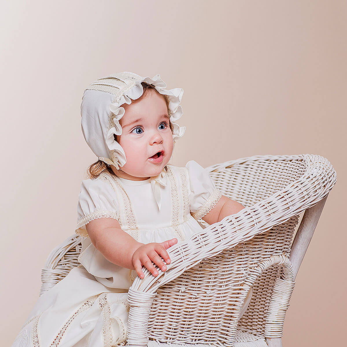 Baby in a white dress and Lace Baptism Bonnet sitting in a wicker chair against a beige background
