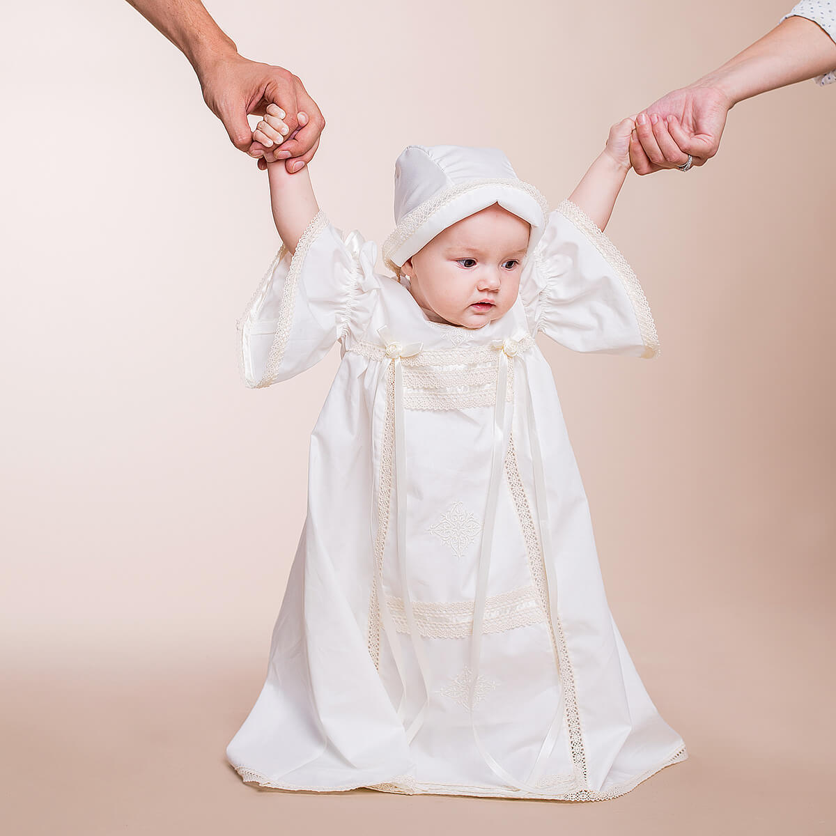 Baby in a Heirloom Christening Dress with ruffled sleeves and a hat, standing between two adult hands on a beige background.