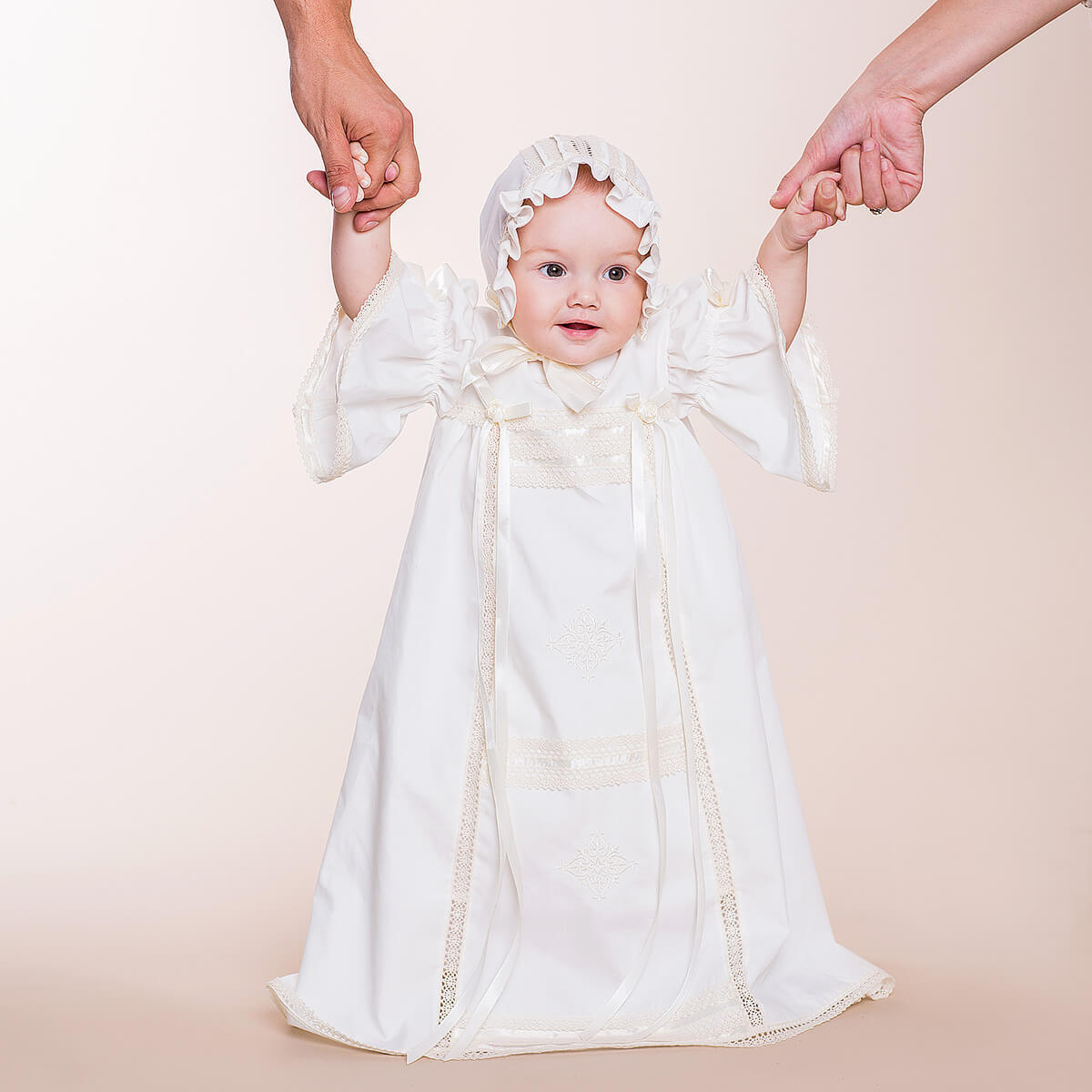 Baby in a Heirloom Christening Dress with a hood being held by two hands against a beige background